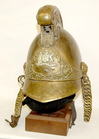 Brass Warrior's Helmet on Wooden Stand: Brass Warrior's Helmet on Wooden Stand: With a dragon crest and side decorations; emblem of a lady, a castle a rearing animal and anchor with "Misi Dominus Frusta"; no makers mark. (Part of lower embl