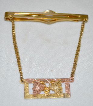 Black Hills Tie Clip With 12K Colored Gold: Black Hills Tie Clip With 12K Colored Gold: the plain gold filled clip has a short chain on both ends. They are connected to a marked Black Hills Gold 12K rectangle with grape bunch and leaves.
