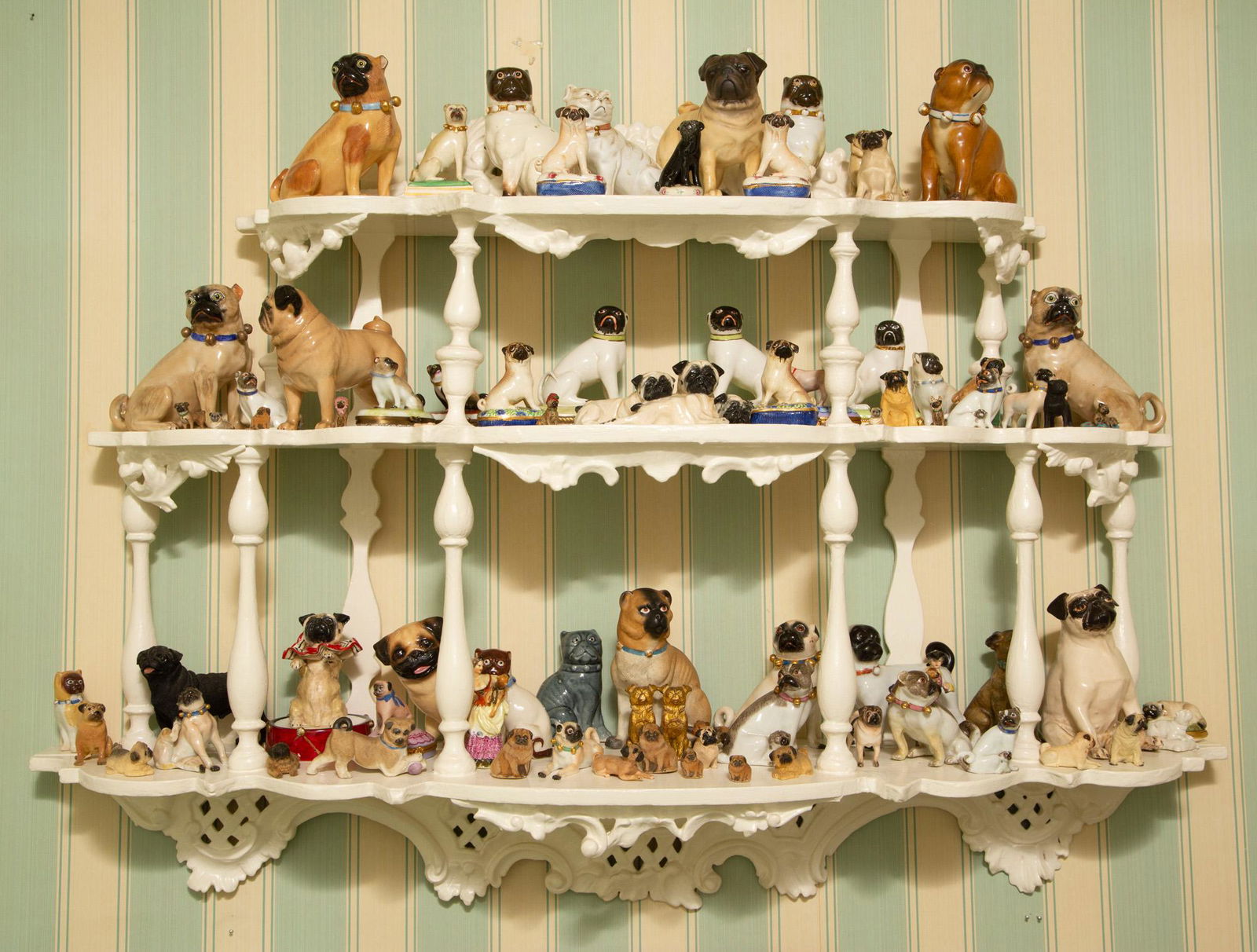 The Pug Installation by Brigid Berlin Comprising a painted wood three-tier wall shelf and: The Pug Installation by Brigid Berlin Comprising a painted wood three-tier wall shelf and approximately seventy figures of pugs in various materials including porcelain. Height overall 27 inches, widt