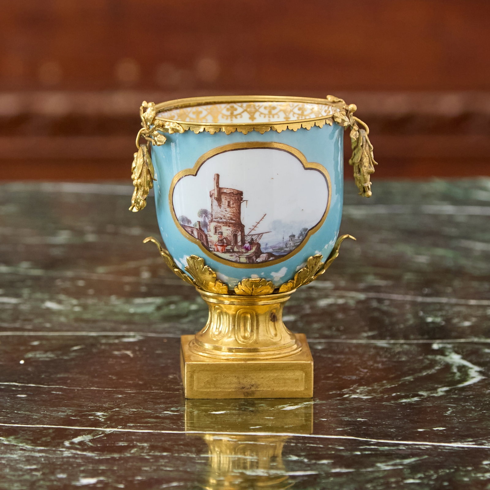 GILT-BRONZE MOUNTED MEISSEN PORCELAIN PALE-BLUE GROUND CUP Circa 1745, the mounts 19th century: Painted front and back with a quatrefoil reserve of either a harbor view or figures in a landscape, the interior enriched with a Gitterwerk band, the metal rim with suspended foliate handles, raised o