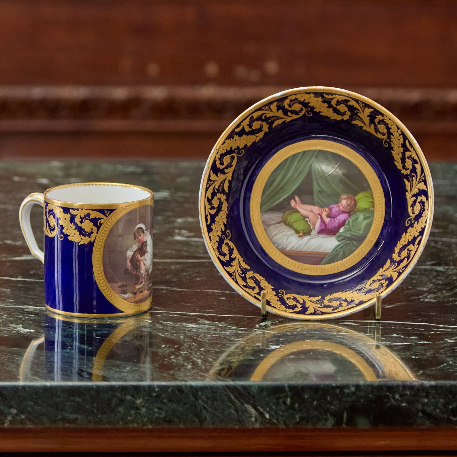 SEVRES PORCELAIN (LATER-DECORATED) COBALT-BLUE GROUND CUP AND SAUCER (GOBELET LITRON) The porcelain: Finely painted after Jean Baptiste Greuze with either a young seated girl in her nightdress and cap clasping a dog or with a young boy asleep in a sumptuous bed, the borders enriched with a foliate Vi