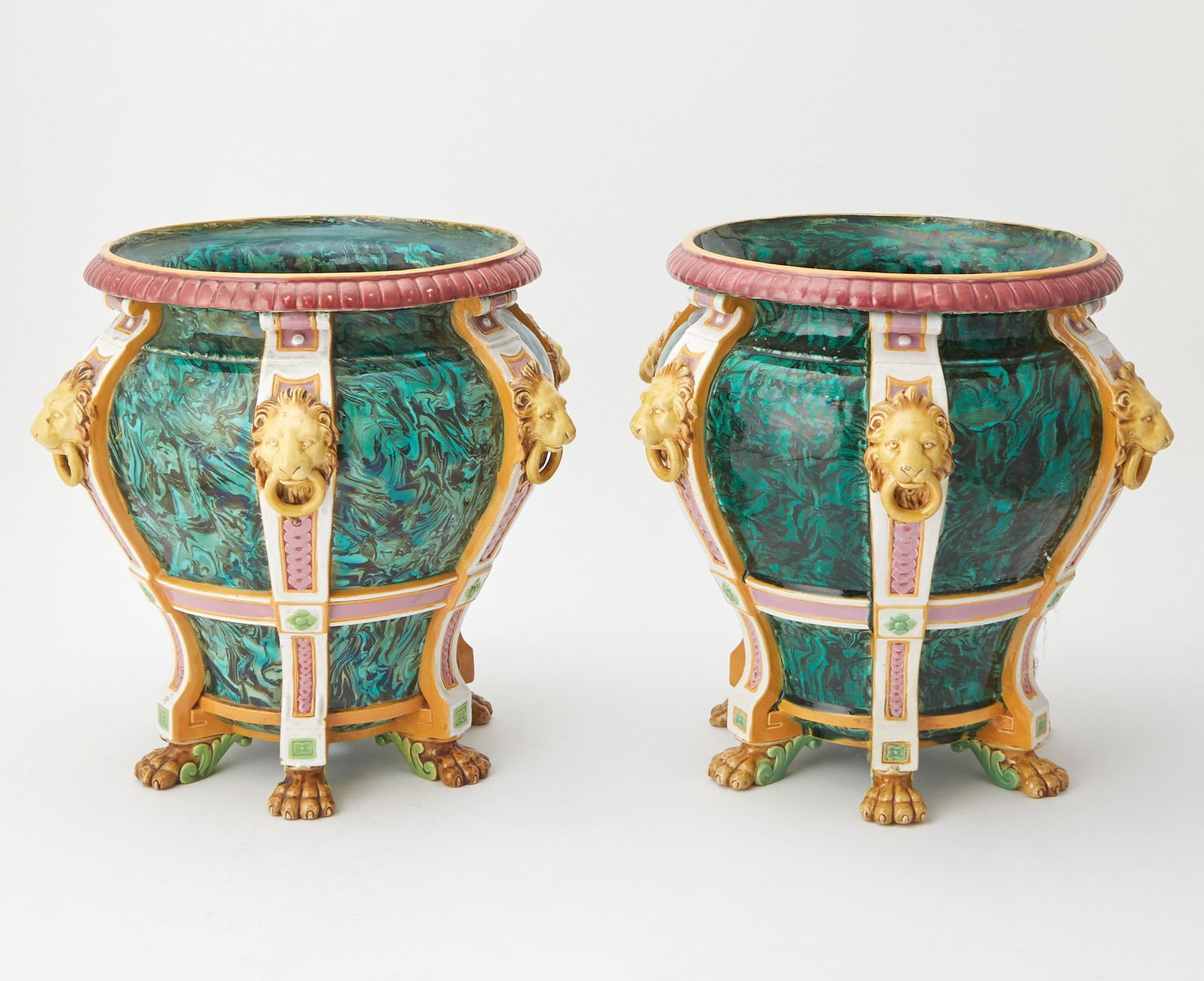 Pair of Minton Style Majolica Faux Malachite-Ground Footed Jardinières: Each of urn form, molded with six white colored vertical straps with pink guilloché bands, golden lion mask and ring handles, with a malachite glaze on both the interior and exterior sides, raised on