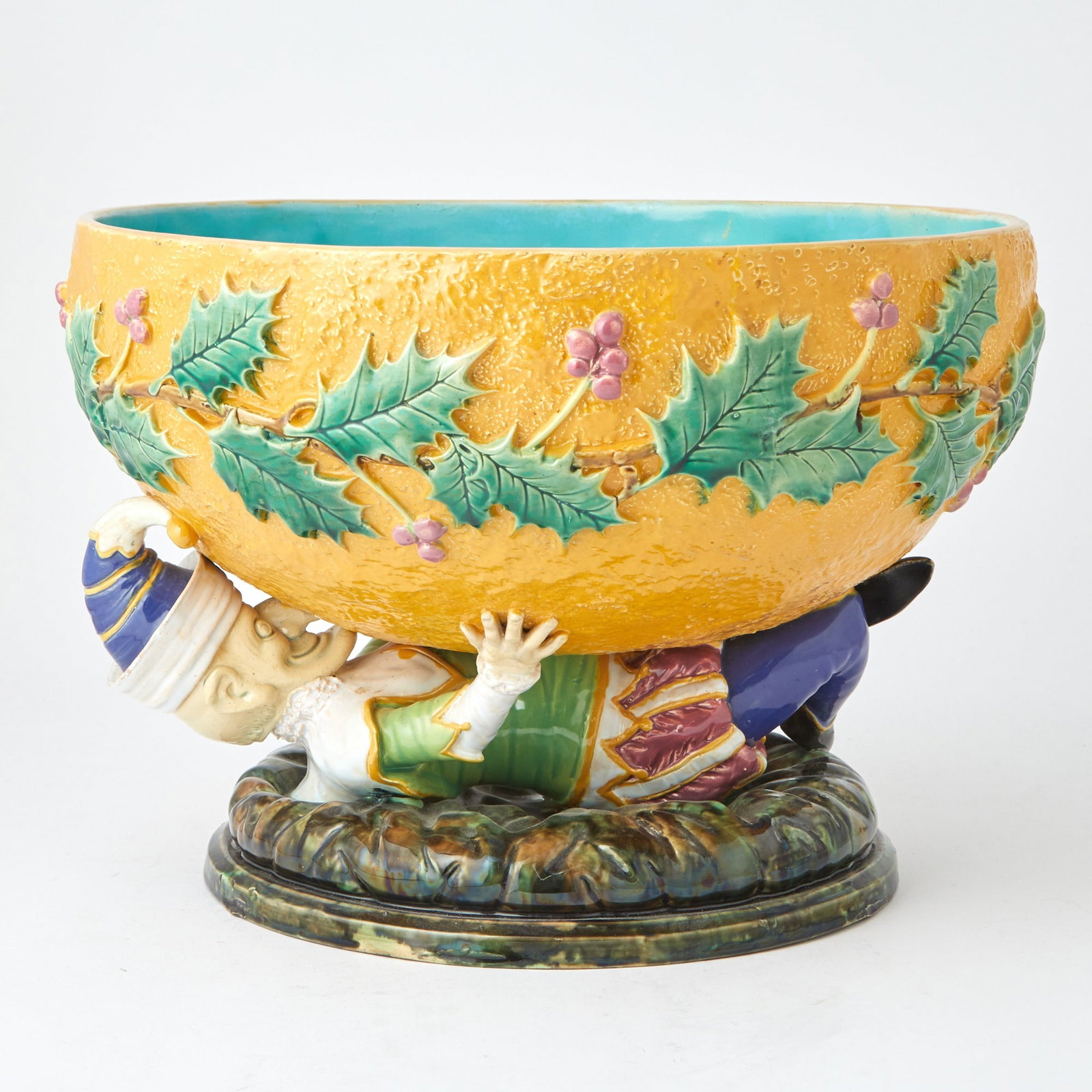 George Jones Majolica Orange-Ground "Mr. Punch" Punch Bowl Circa 1875: Modelled as a figure of Punch, dressed in a brightly coloured jester costume, lying on his back on the base and holding a bowl modelled as an orange peel circled by holly leaves and berries, the insid
