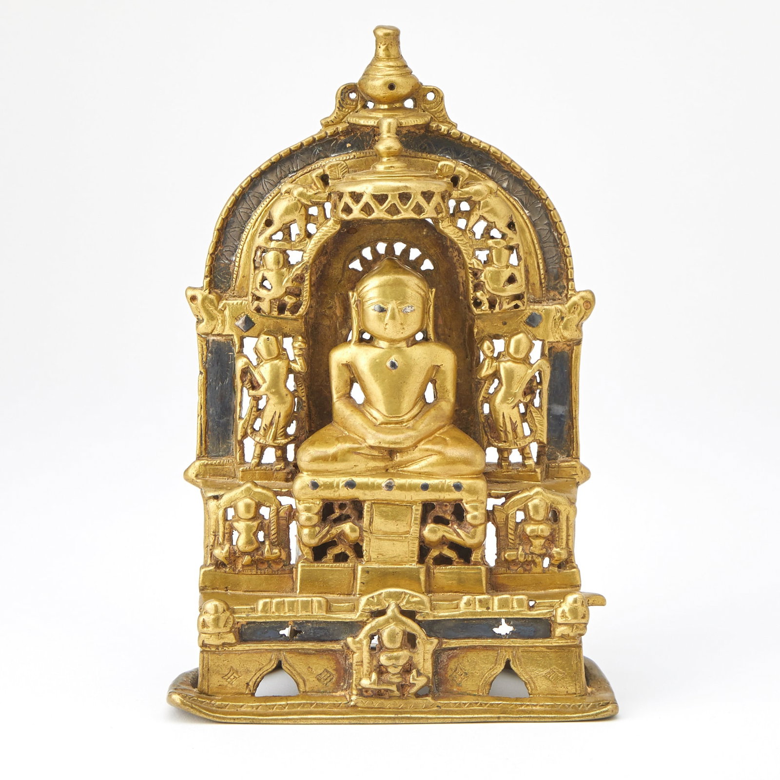 An Indian Silver-Inlaid Bronze Jain Votive Dated 1517: The parcel gilt bronze shrine cast to depict the central figure of Jina with silver eyes and seated on a throne surrounded by an openwork arch with attending devas; inscription verso. (8 inches tall, 