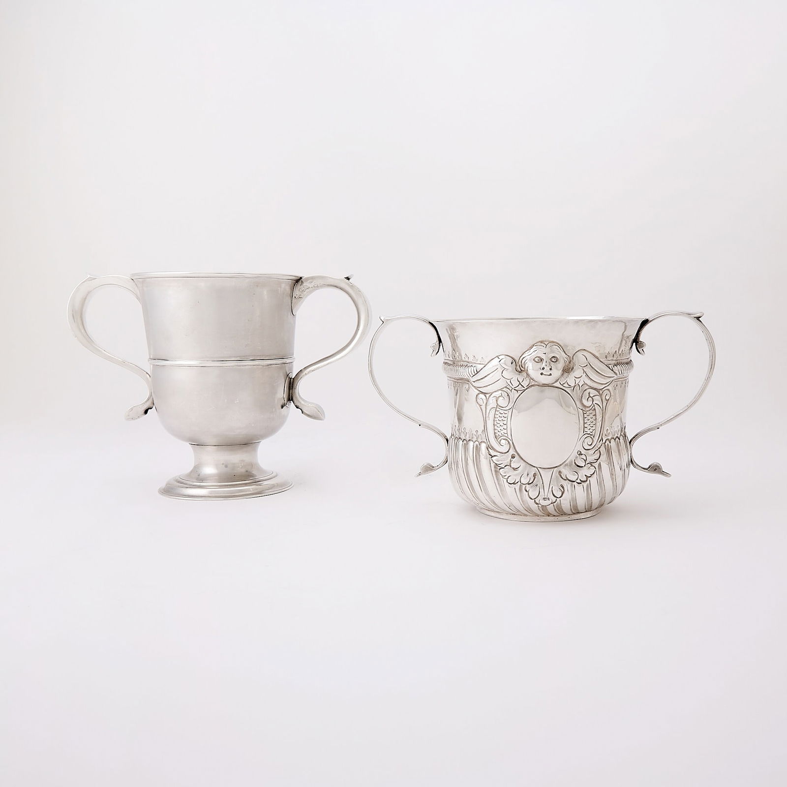 Two George I/II Sterling Silver Two Handled Cups: Comprising a George I example with spiral fluting and cartouche marked James Smith, London, 1722, height 4 3/4 inches (11.5 cm), width over handles 8 1/4 inches (21 cm); the other George II example ma