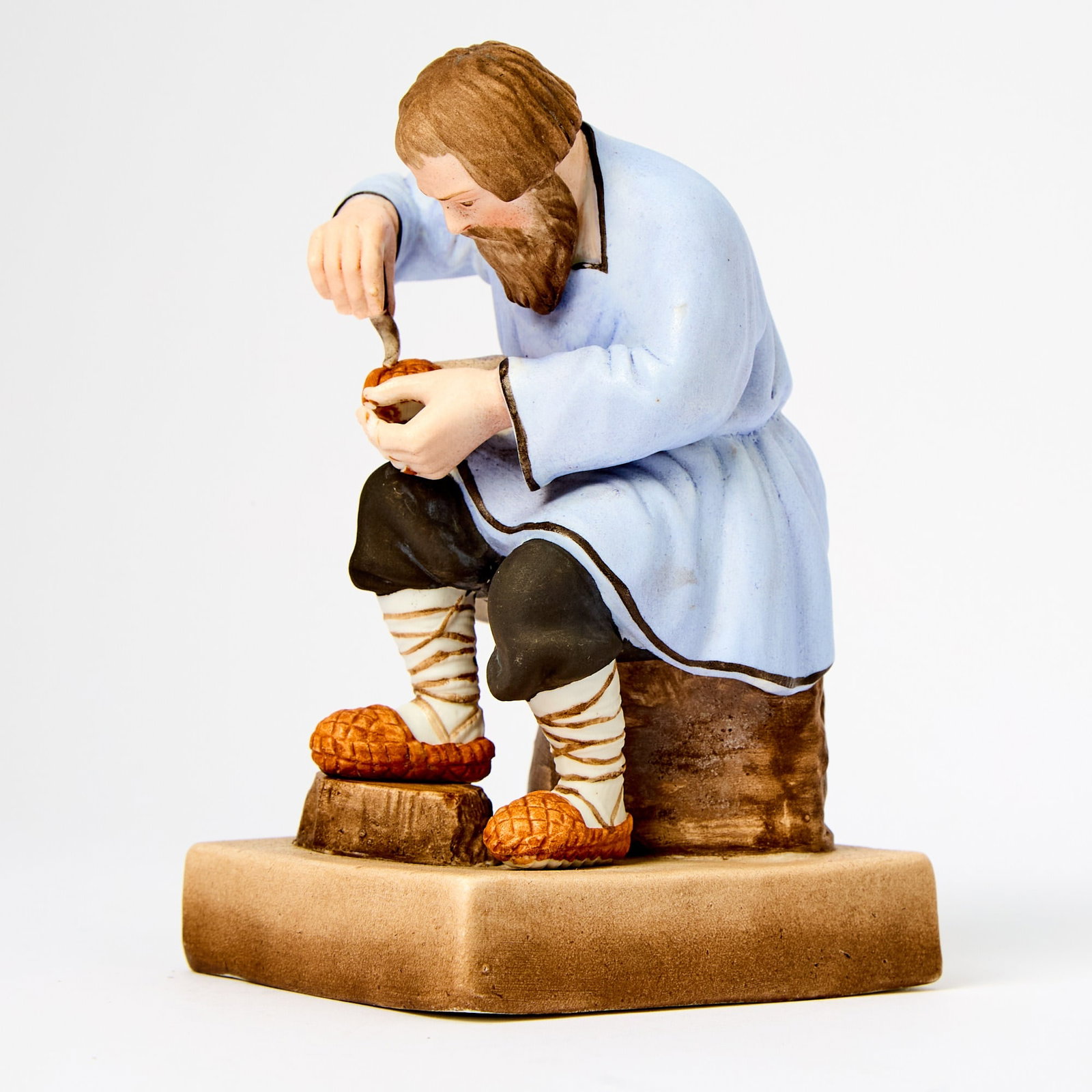 Russian Porcelain Figure of a Bast Shoemaker: Gardner Manufactory, Verbilki, 1870-1890 Modeled as a bearded man wearing a blue rubakha, black pants and bast shoes, seated on a tree trunk, with one foot raised on a stump, on a square simulated woo