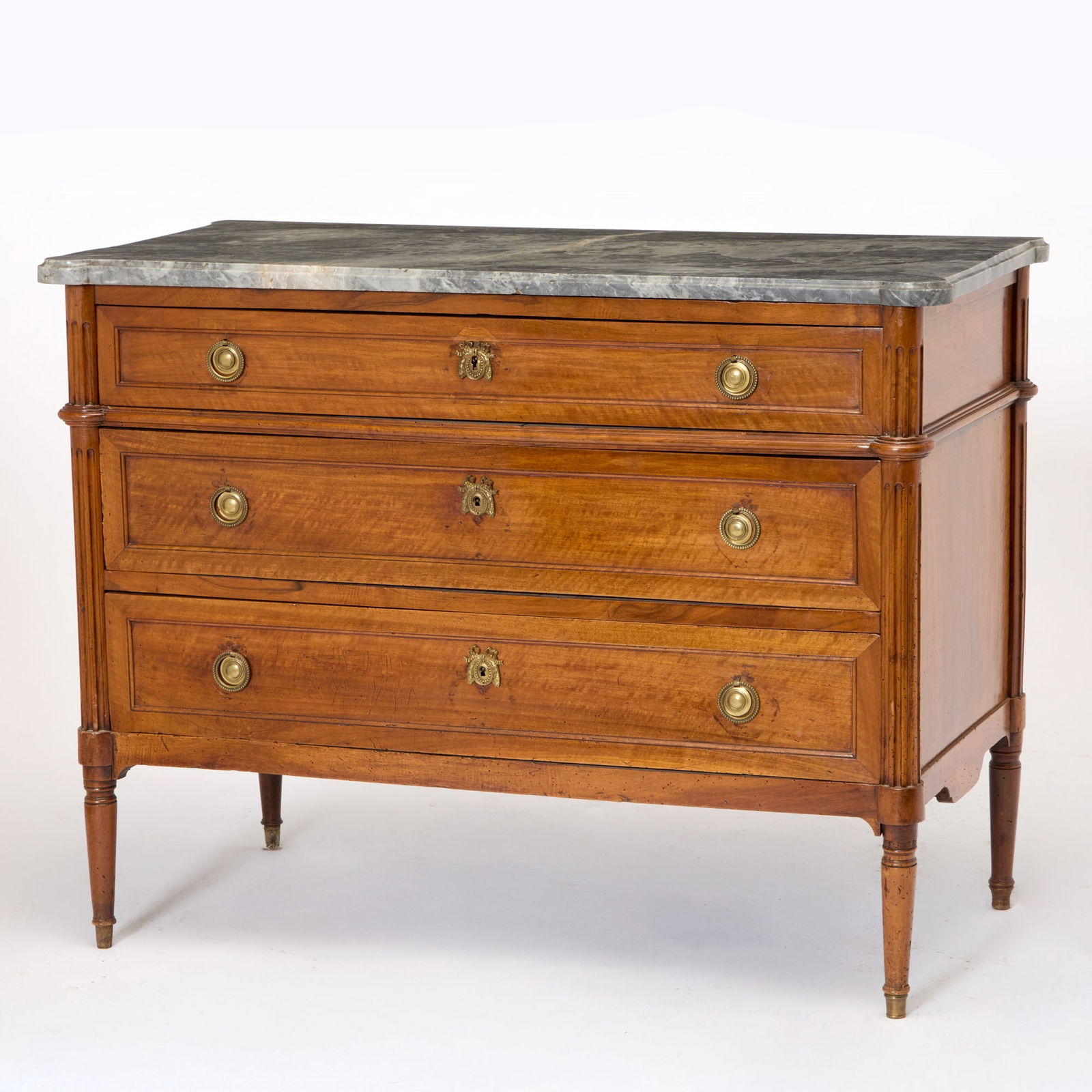 Louis XVI Fruitwood Commode: Late 18th century The molded rectangular white veined grey marble top with rounded corners above a conforming case fitted with three long drawers flanked by fluted standards continuing to tapering fee