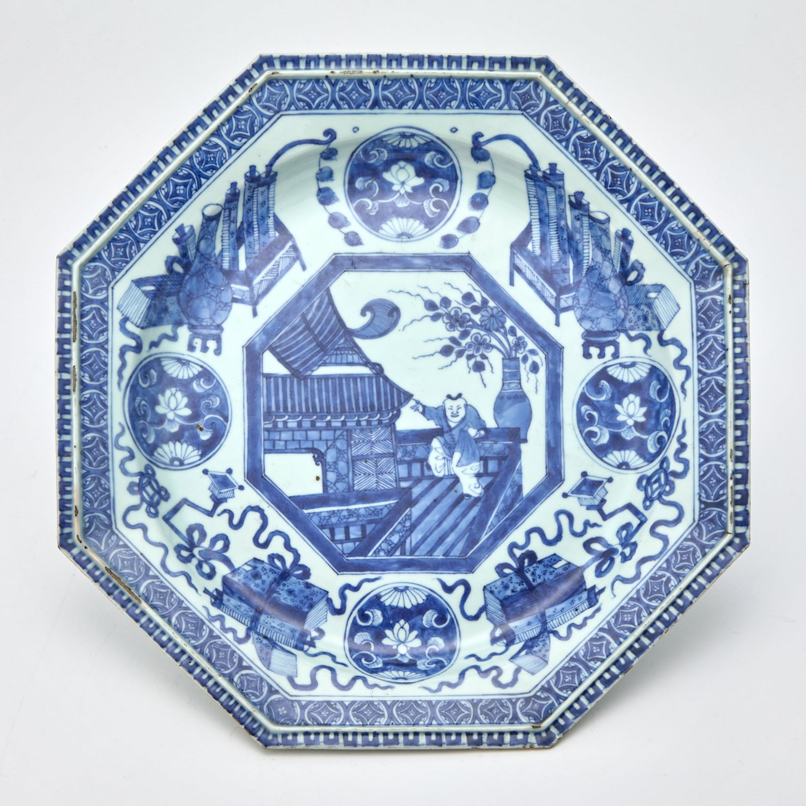 A Chinese Blue and White Porcelain Octagonal Dish Kangxi Period: The shallow, rounded sides rising to an everted rim with a raised rib to inner rim; decorated to show an octagonal panel depicting a boy in an architectural setting, encircled by scholar's objects, lo
