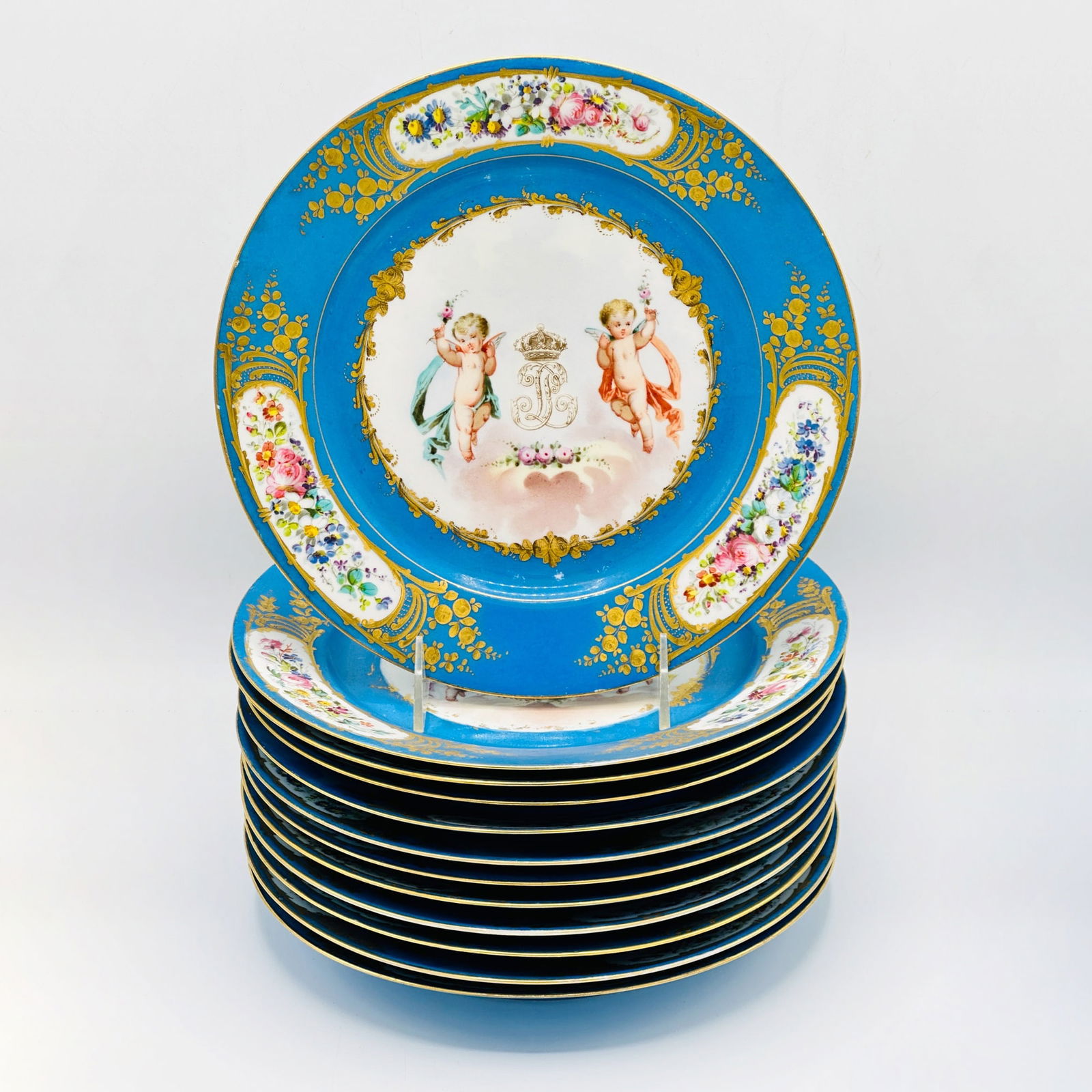 Set of Twelve Sevres Porcelain Dinner Plates: Late 19th centuryEach with blue rim with gilt highlights and floral reserves, centering cherubs flanking a crown.Diameter 9 3/4 inches.