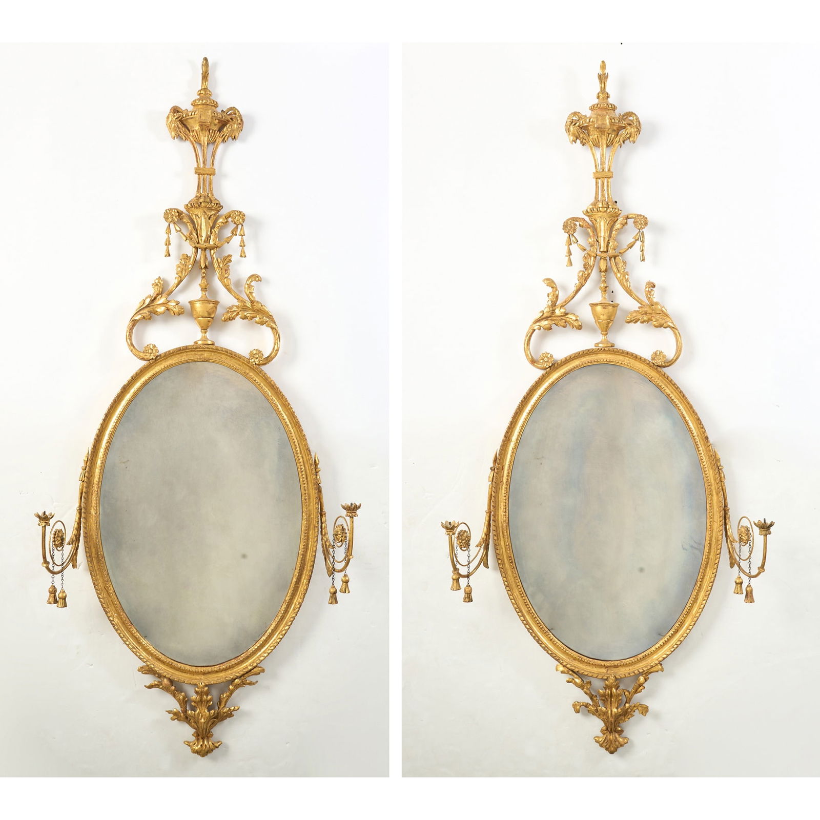 Pair of George III Giltwood Oval Girandoles: Circa 1770 Each later oval mirror plate within a molded surround surmounting an acanthus carved pediment flanking a tall urn centered by husk, swags and floral rosettes, issuing two scrolling candle a