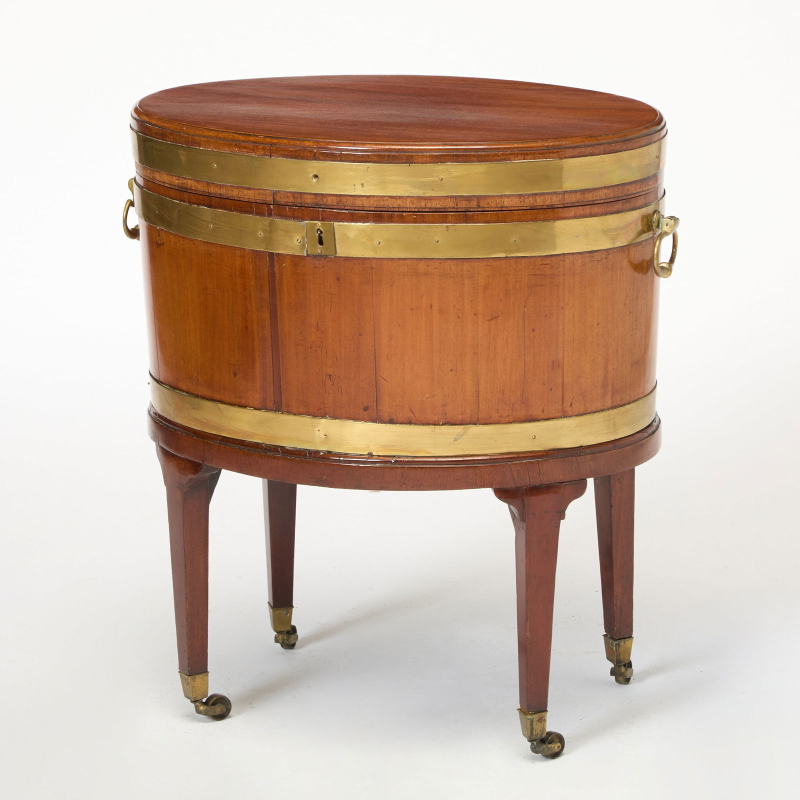 George III Brass-Mounted Mahogany Wine Cooler: Early 19th century The oval hinged top above a conforming tapering case enclosing a green baize-lined well, with applied brass handles, on a later stand with tapering square legs and brass casters. He