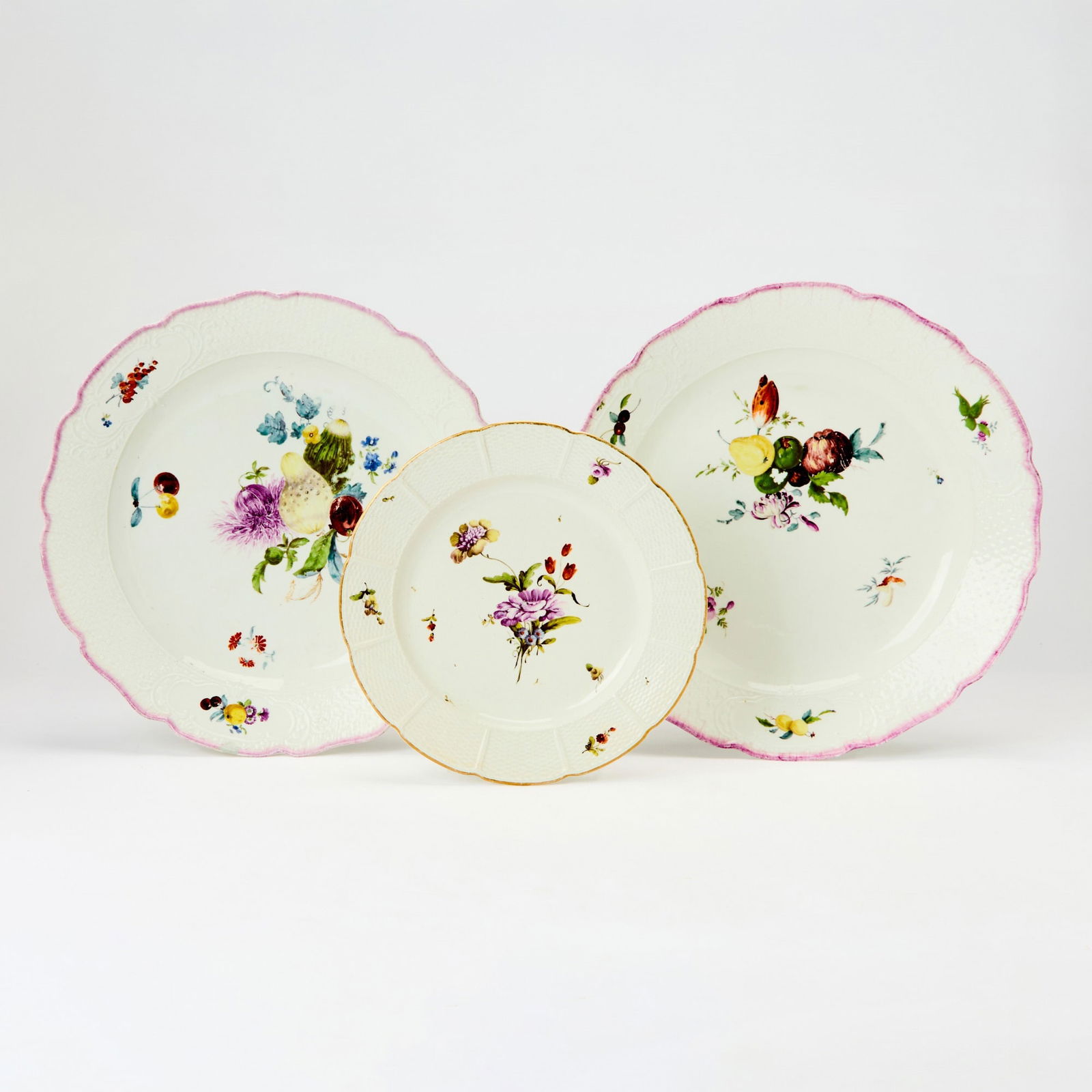 Two Meissen Porcelain Chargers and a Ludwigsburg Porcelain Plate: Mid-18th century Each with a moulded and basket-weave textured border heightened with a purple rim and a gilt rim, painted in colors with a delicate arrangement of fruits and flowers. Diameter Meissen