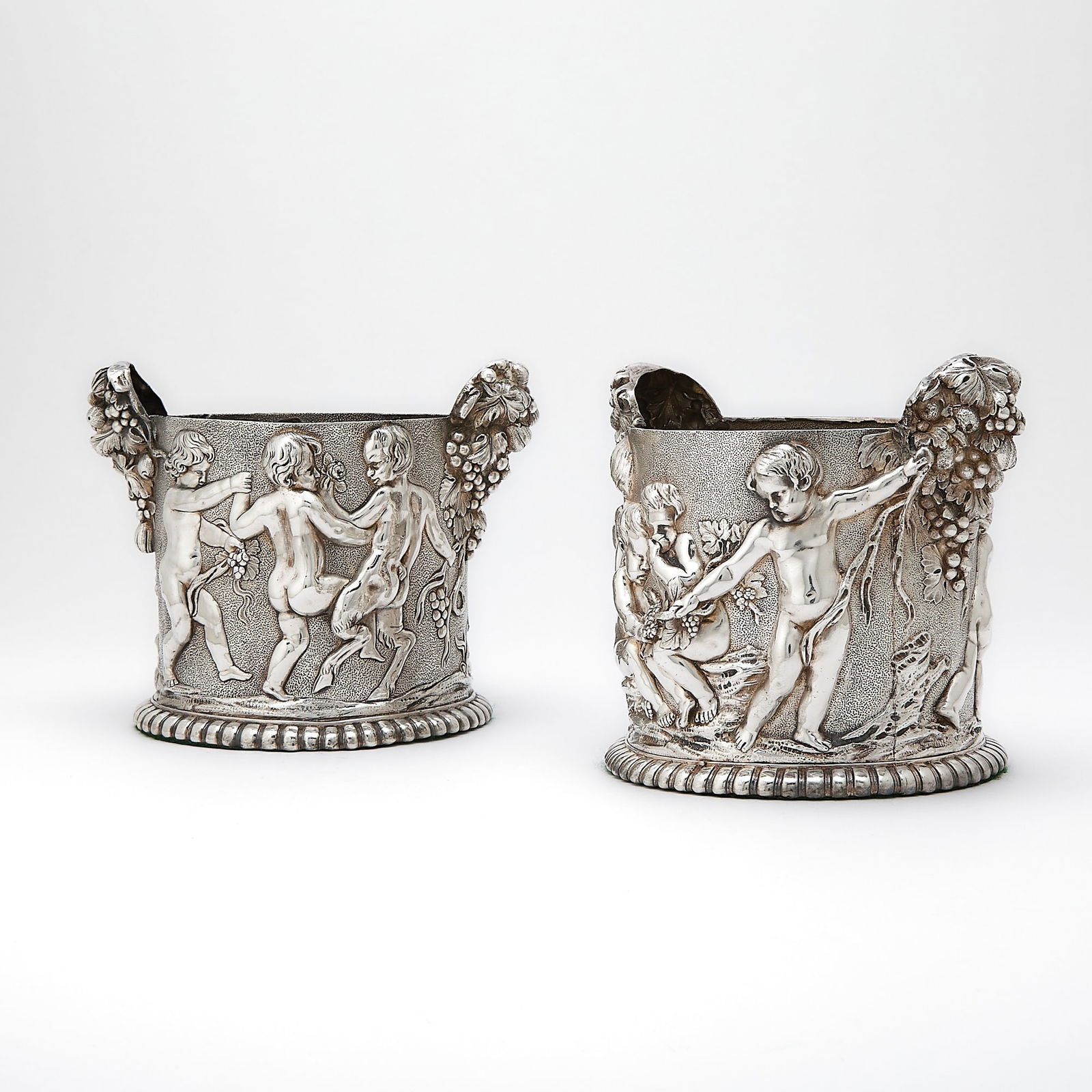 Pair of Sterling Silver Bottle Holders: Maker's mark RC, probably for Richard Comyns, late 20th century Each cylindrical with scenes of putti, the handles of grape clusters, on turned wooden base. Height over handles 6 inches (15 cm), lengt
