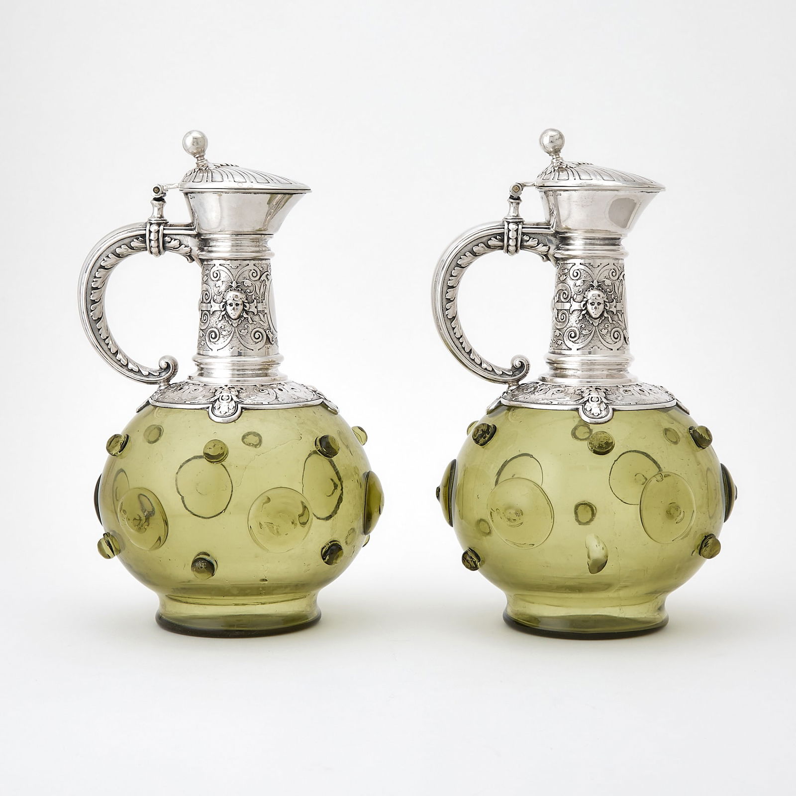 Pair of Continental Silver Mounted Green Glass Claret Jugs: Late 19th century The studded globular bodies with slender necks with masks and scrolls, with hinged covers and loop handles. Height 10 1/2 inches (26.5 cm), weighted.