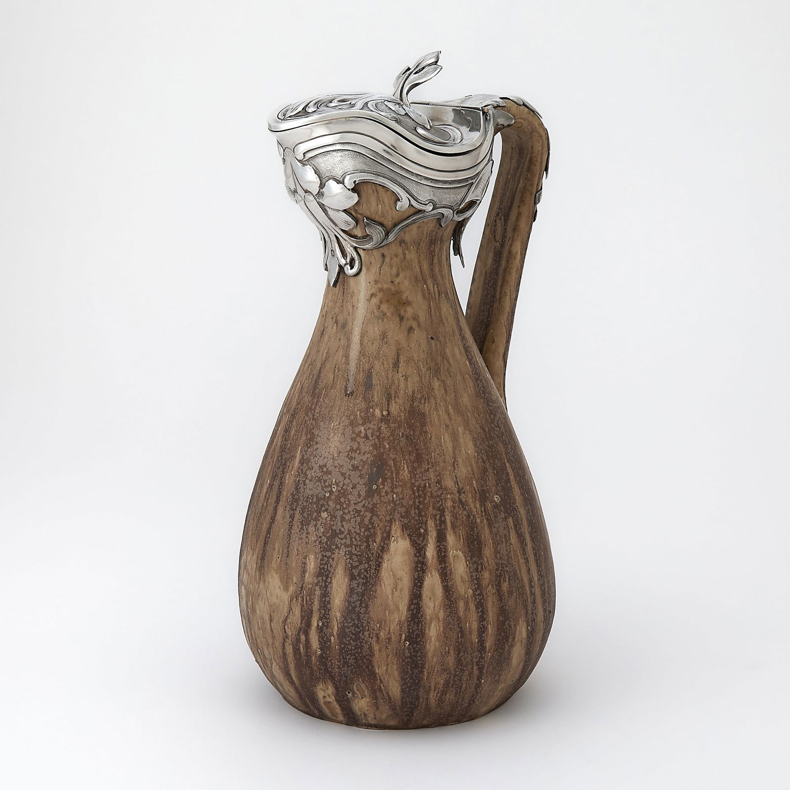 French Art Nouveau Silver Mounted Glazed Ceramic Ewer: Late 19th/ early 20th century The ovoid body with raised loop handles, the neck mount with stylized foliate scrolls, the hinged cover with foliate thumbpiece. Height 10 1/2 inches (26.5 cm),