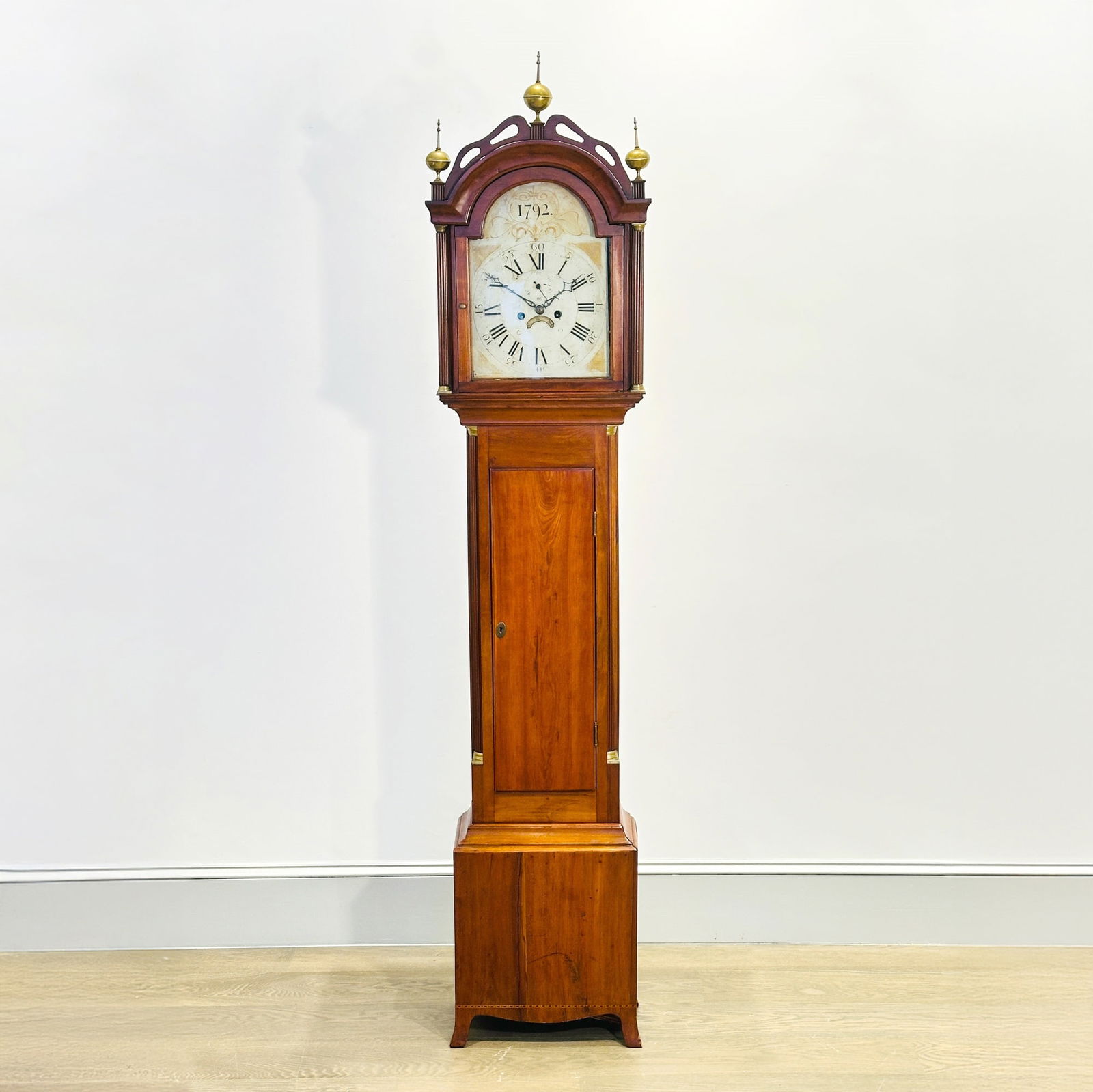 Federal Inlaid Cherry Tall Clock: Massachusetts or New Hampshire, Early 19th Century The hood with glazed tombstone door and white-painted iron dial dated 1792, above a waist with fluted quarter columns and base with flaring French