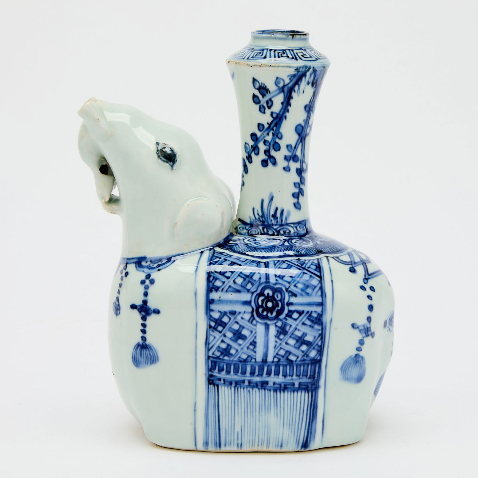 A Chinese Blue and White Porcelain Elephant-Form Kendi Late Ming Dynasty: Modeled in the form of a recumbent elephant with its head uplifted, the trunk issuing as the spout; the tubular neck encircled by bands of key-fret and scrolling motifs framing budding branches; the b