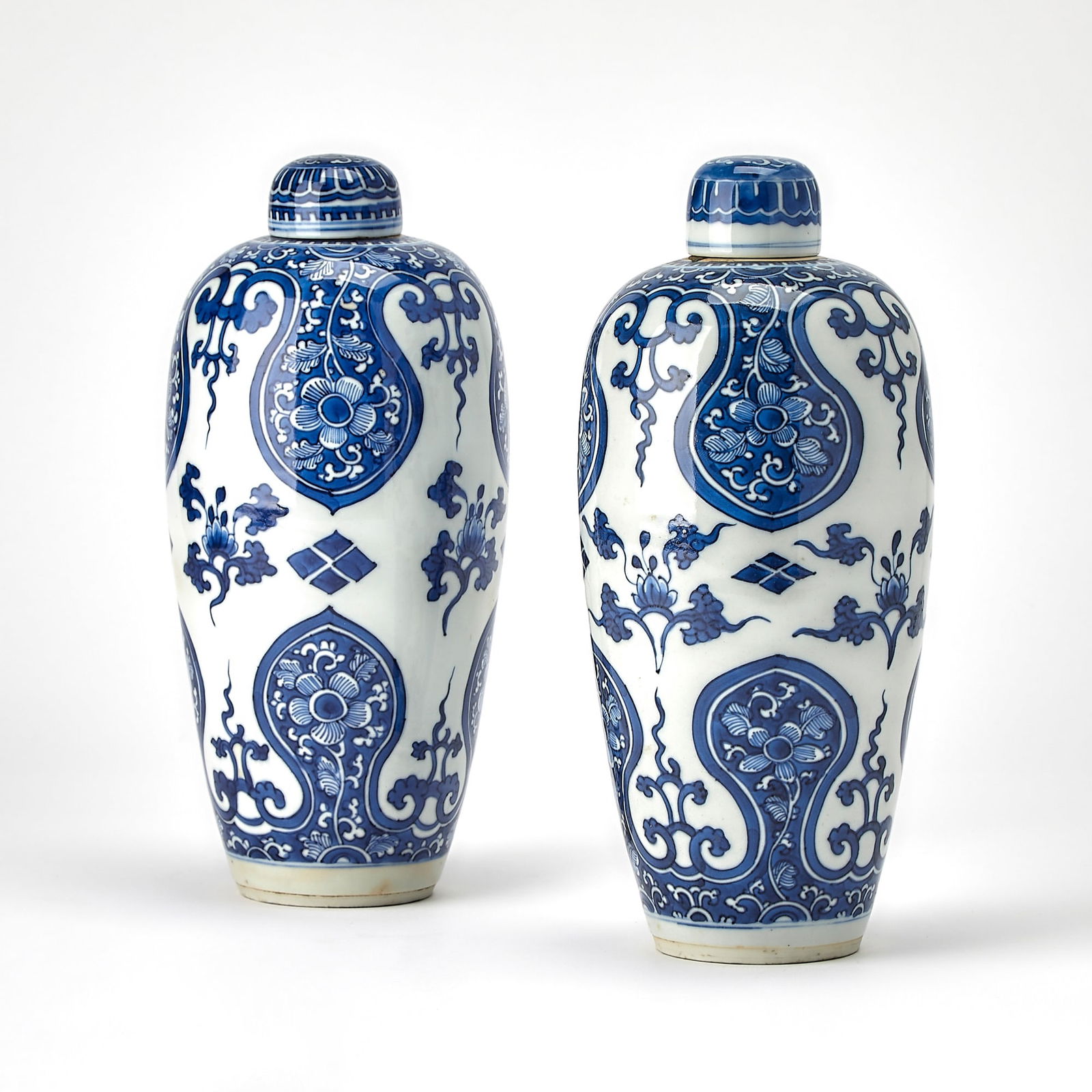 A Pair of Chinese Blue and White Porcelain Vases and Covers Kangxi Period: Each of ovoid form with a domed cover; decorated with floral sprays and diaper patterns, framed by large stylized foliate-shaped cartouches enclosing blossoms borne on leafy vines. (Height 10 inches)