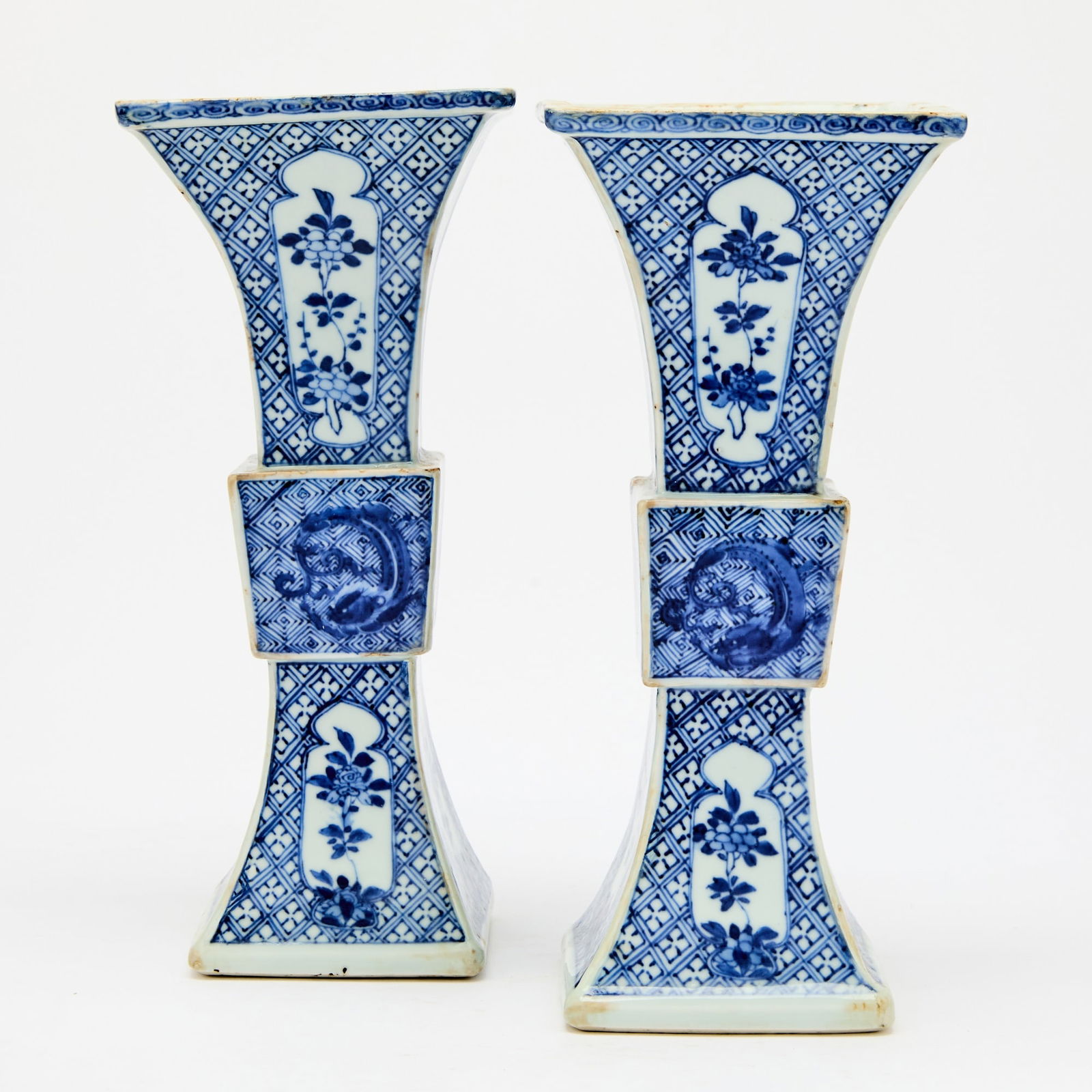 A Pair of Chinese Blue and White Porcelain Gu Vases Kangxi Period: Each of square form, the upper and lower sections decorated with floral sprays within shaped cartouches reserved on a diaper ground, the mid-section with coiling chilongs over a key-fret ground. (Heig