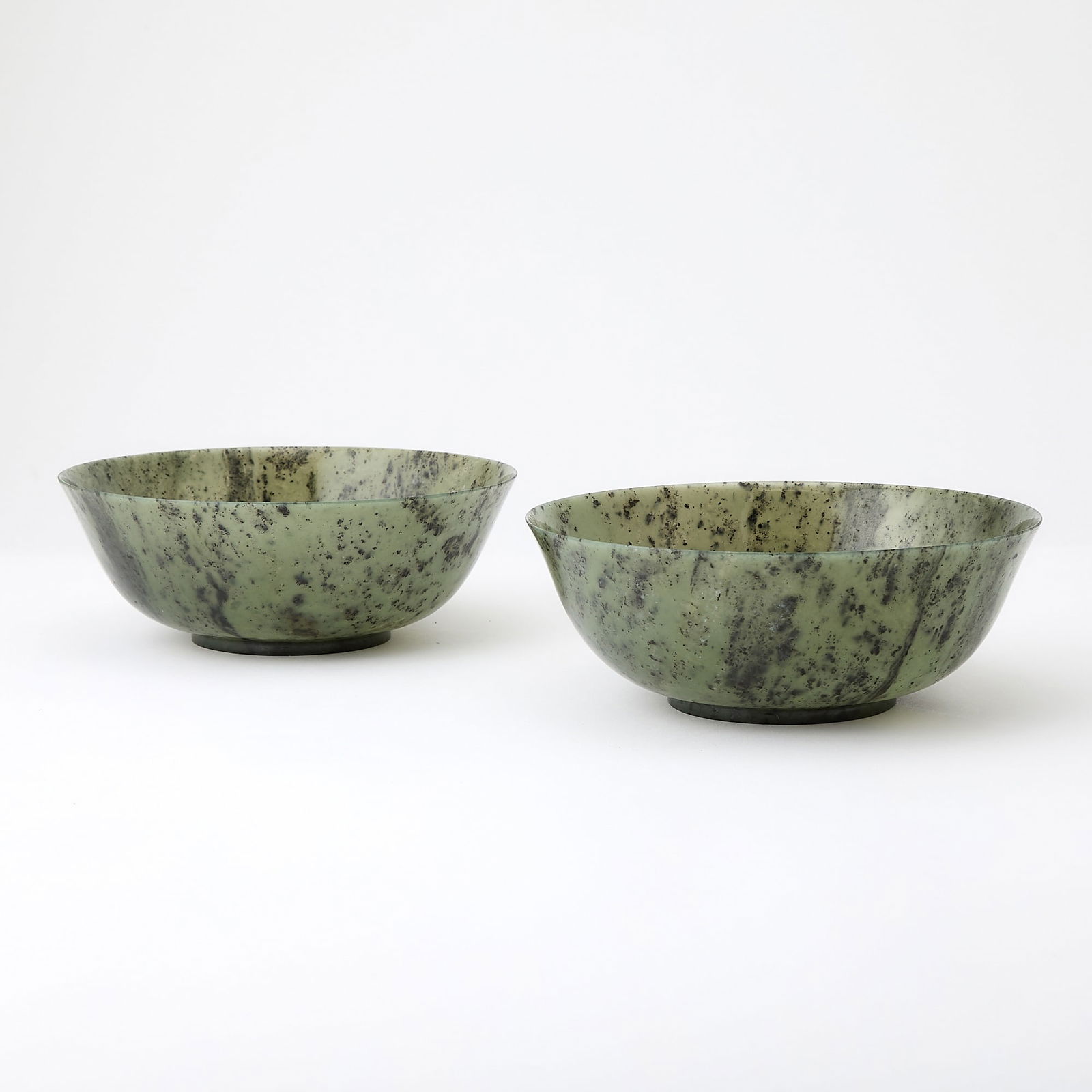 A Pair of Chinese Spinach Jade Bowls 19th century: Each thinly carved with rounded sides rising to a slightly everted rim and resting on a short foot ring; the jade of olive-green tone flecked with dark green. (Diameter 7 1/4 inches)