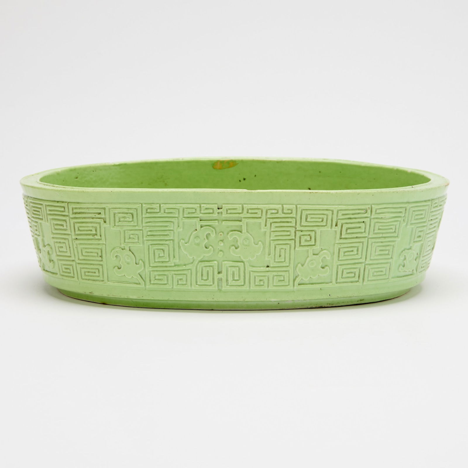 A Chinese Carved Lime Green Porcelain Narcissus Bowl 19th Century: Of oval form and tapering slightly to foot; decorated to exterior with relief carved geometric pattern incorporating chilong heads; the recessed top rim carved in intricate wave pattern. (Width 9 3/4