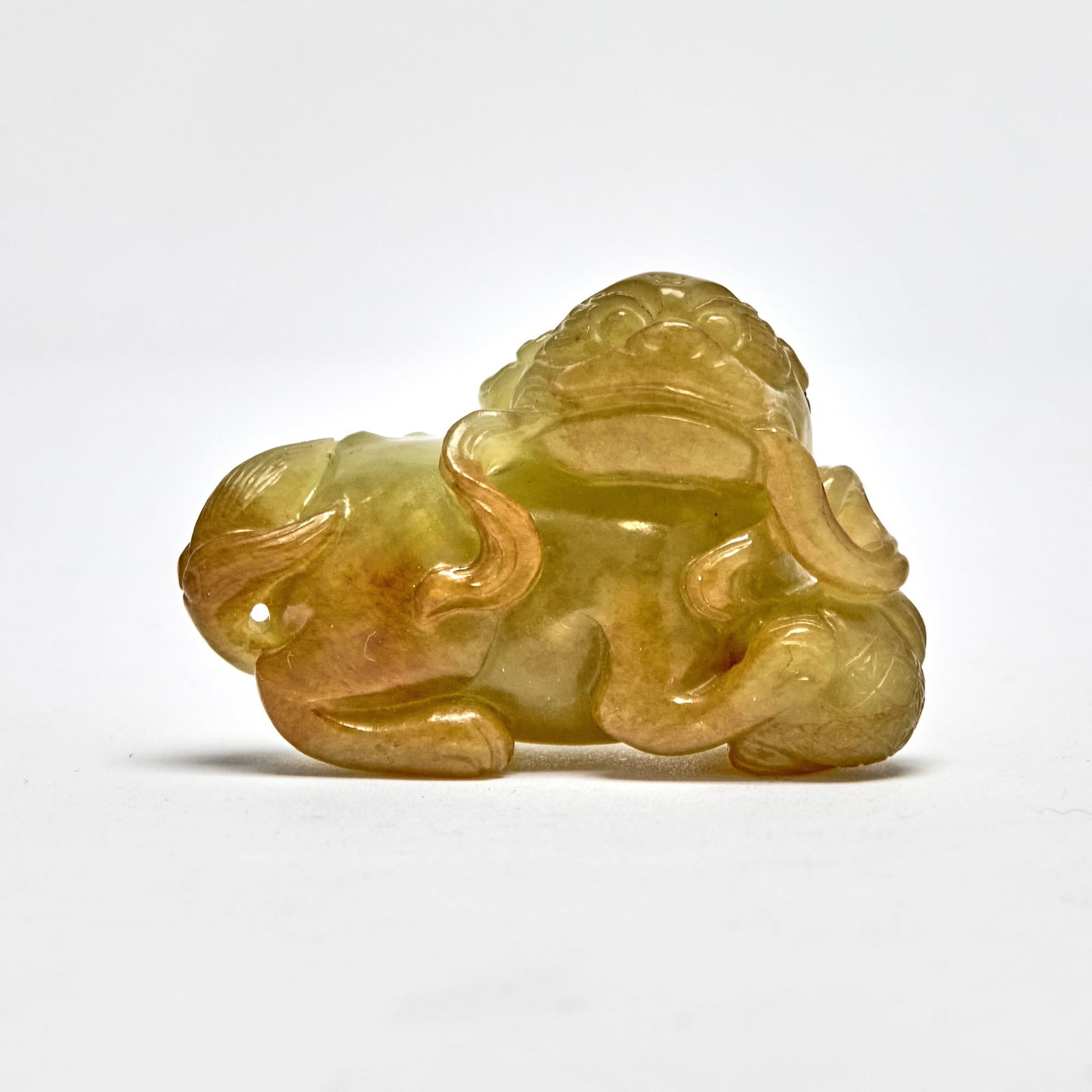 A Chinese Celadon Yellow Jade Carving of a Fu Lion: Carved as a recumbent figure holding a band tying a brocade ball beneath its forepaws. (Length 2 1/4 inches)