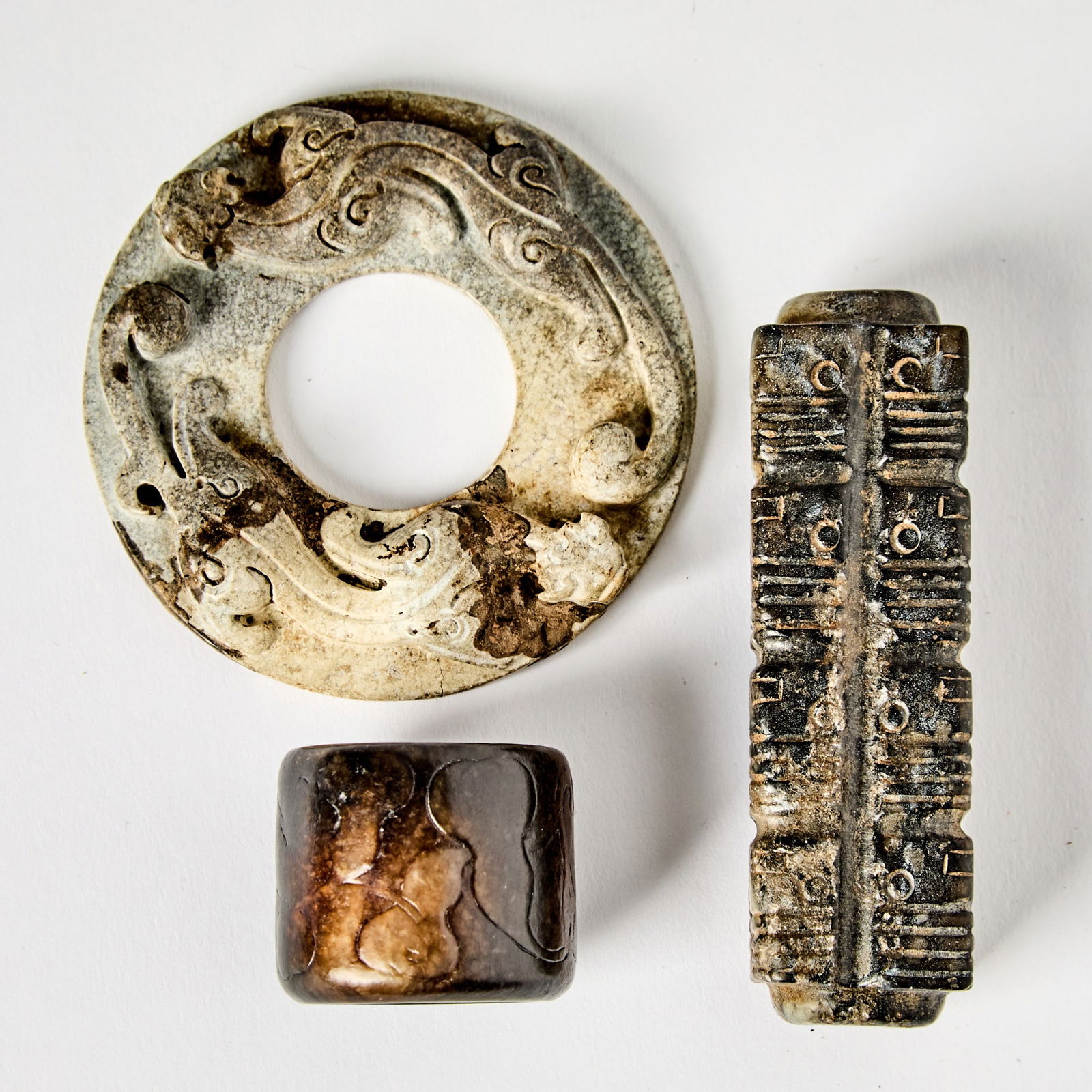 A Group of Three Chinese Archaistic Jade Carvings Ming Dynasty and earlier: Comprising a calcified bi dis with relief qilong, a thumb ring, and a mottled jade cong. (Length of largest 2 3/4 inches)