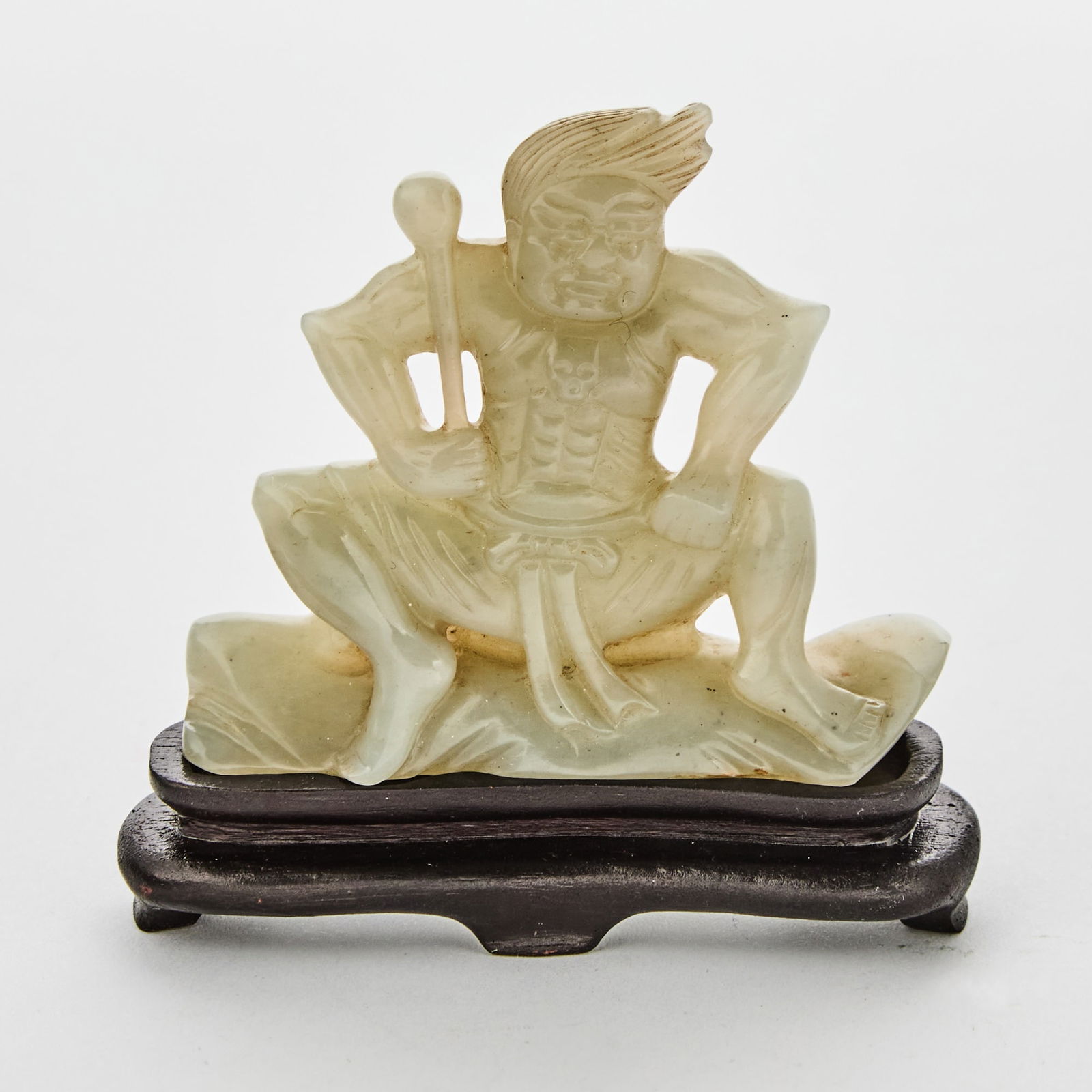 A Chinese Celadon Jade Figural Carving of Vajrapani 17th Century: Carved as a seated figure with a fierce expression, resting both hands on his knees, one hand holding his attribute; with a fitted wood stand. (Height 2 1/4 inches)