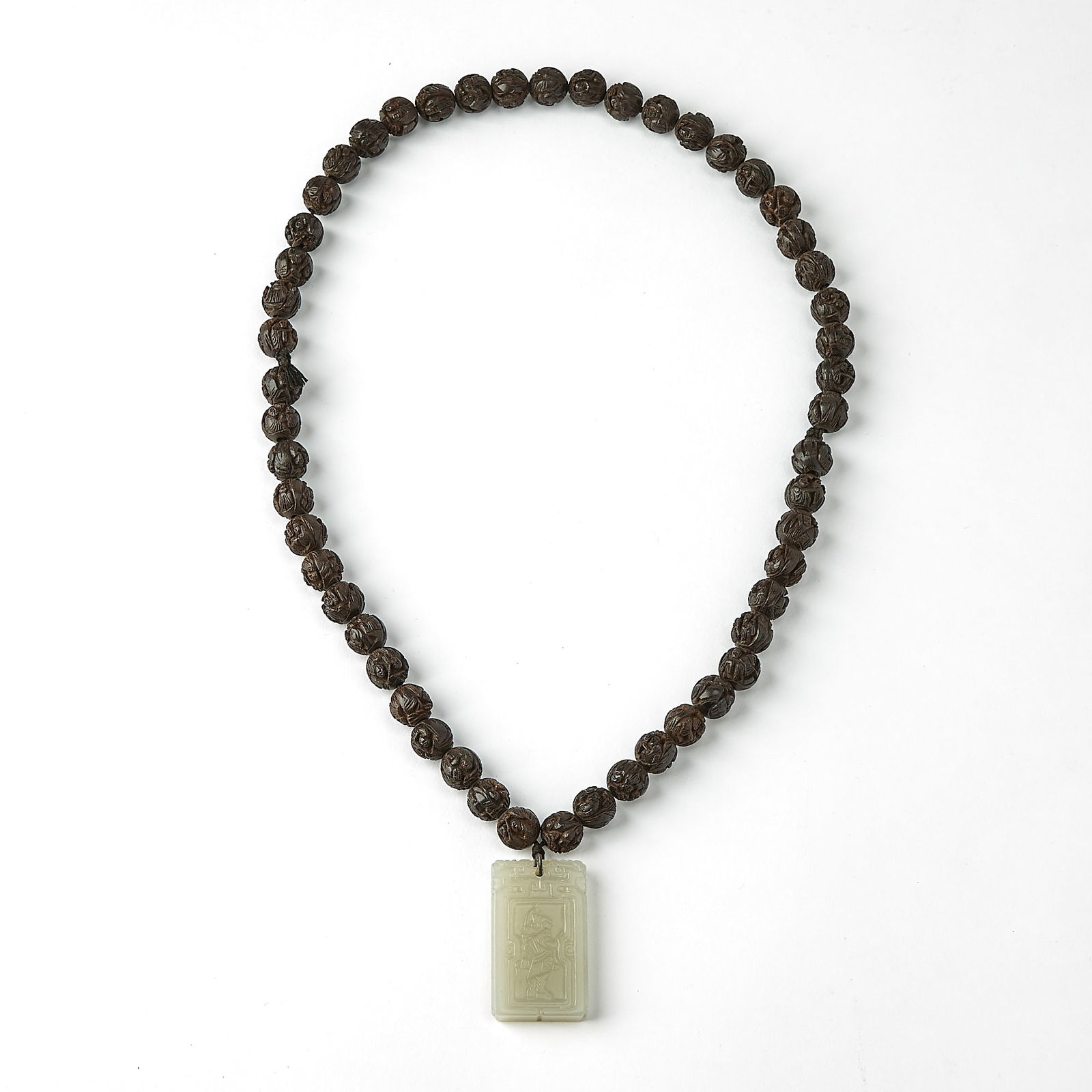 A Chinese Carved Walnut Necklace with a White Jade Pendant Qing Dynasty: The strand of carved beads suspending a rectangular plaque carved in low relief with a standing figure within a shaped cartouche, the reverse carved with a verse and the seals ‘wen’ and &#