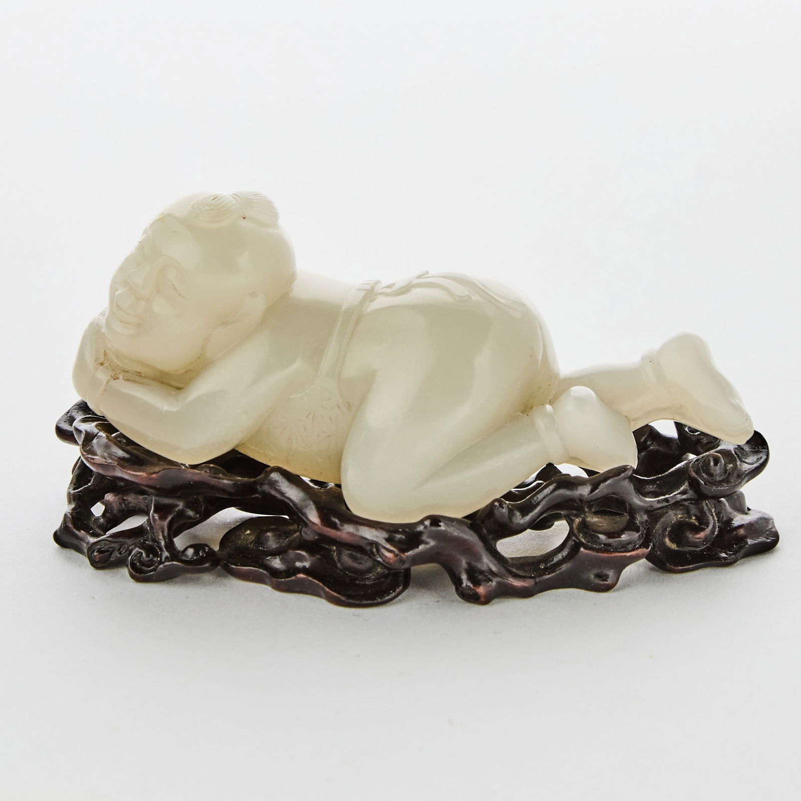 A Chinese White Jade Figural Carving of a Boy: Carved in a prone position holding a ball in his hands, the face bearing a pleasant expression; with a finely carved wood stand. (Length 3 1/2 inches)
