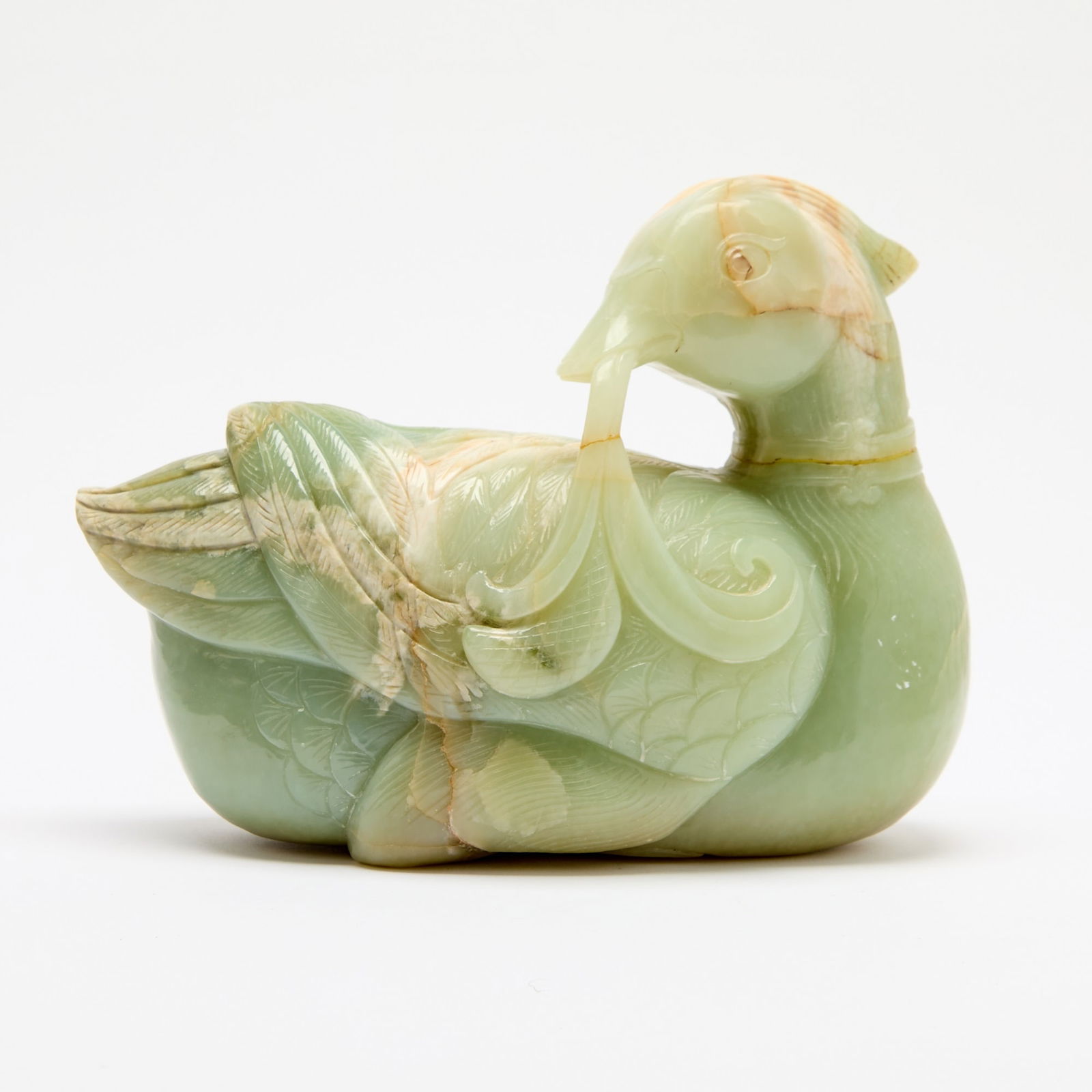 A Large Chinese Celadon Jade Figure of a Duck Republic Period: Carved in the form of a recumbent duck with its head turned back and gazing over its shoulder, holding a leafy spray in its beak; the stone of mottled tone. (Length 9 1/2 inches)