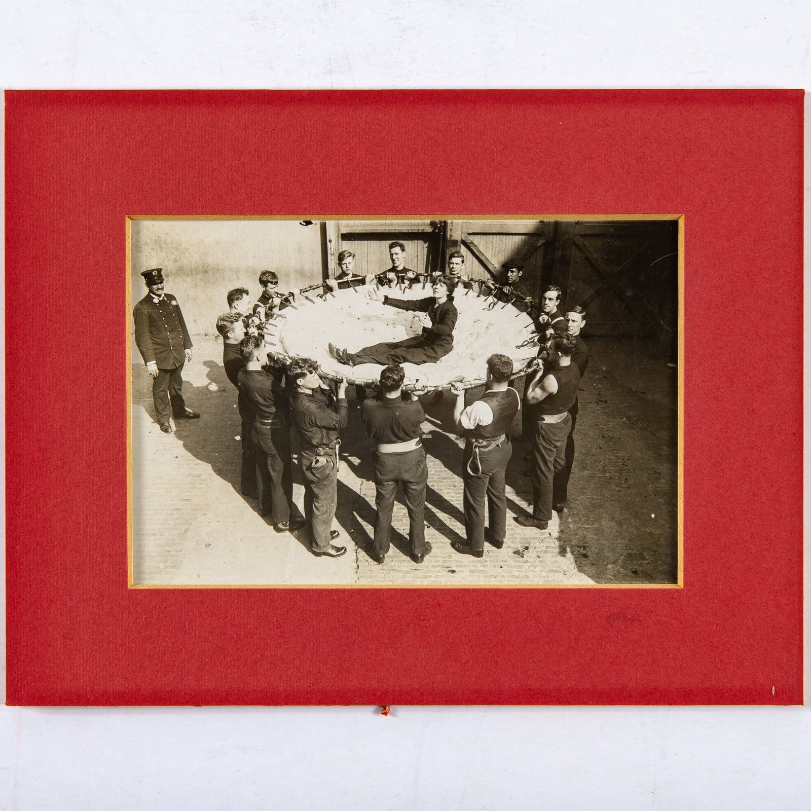 Vintage photograph of Djuna Barnes rescued by firemen: [BARNES, DJUNA] Gelatin silver print of Djuna Barnes on a fireman's safety net, held by a dozen or so firemen, 1914, vintage print. 3 3/4 x 5 7/8 inches (9.5 x 15 cm); dated on verso in ink in a perio