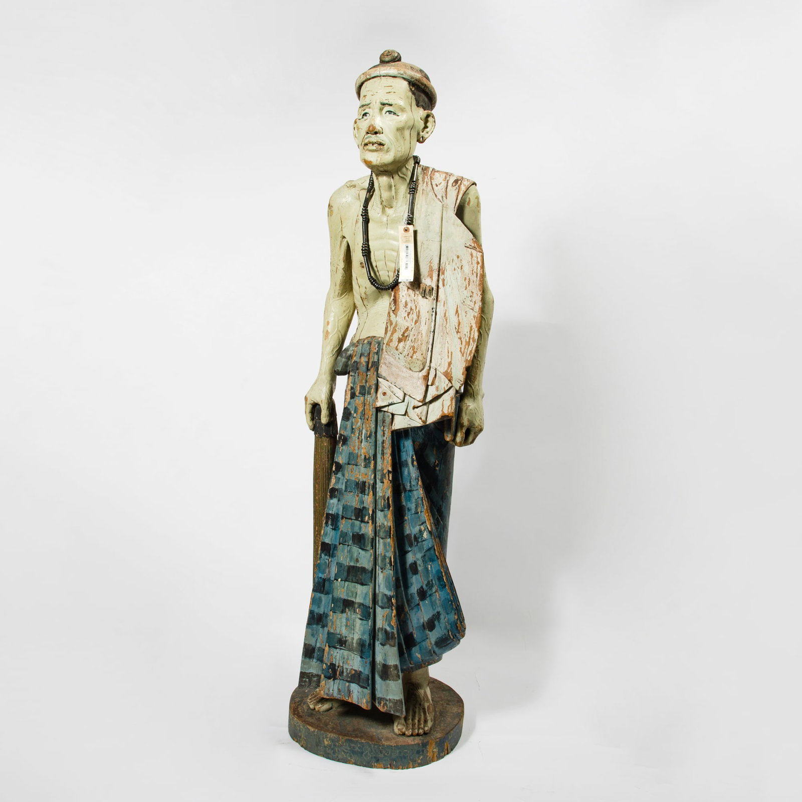 Burmese Painted Wood Figure of a Man 19th-20th Century: The elderly figure supporting himself with a cane, carved with a lean physique and deeply lined face; the figure and his traditional attire decorated with polychrome pigments. (Height 66 inches) Purch