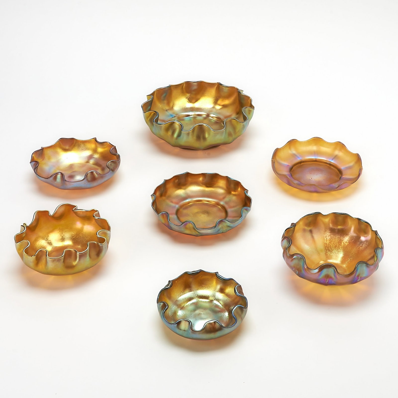 Group of Seven Tiffany Gold Favrile Glass Small Bowls and Underplates: Each with a crimped rim, in transparent amber colored glass, gold iridized, six examples engraved L.C.T., one example engraved X307 L.C. Tiffany-Favrile. Height of largest 2 1/4 inches, width 6 5/8 in
