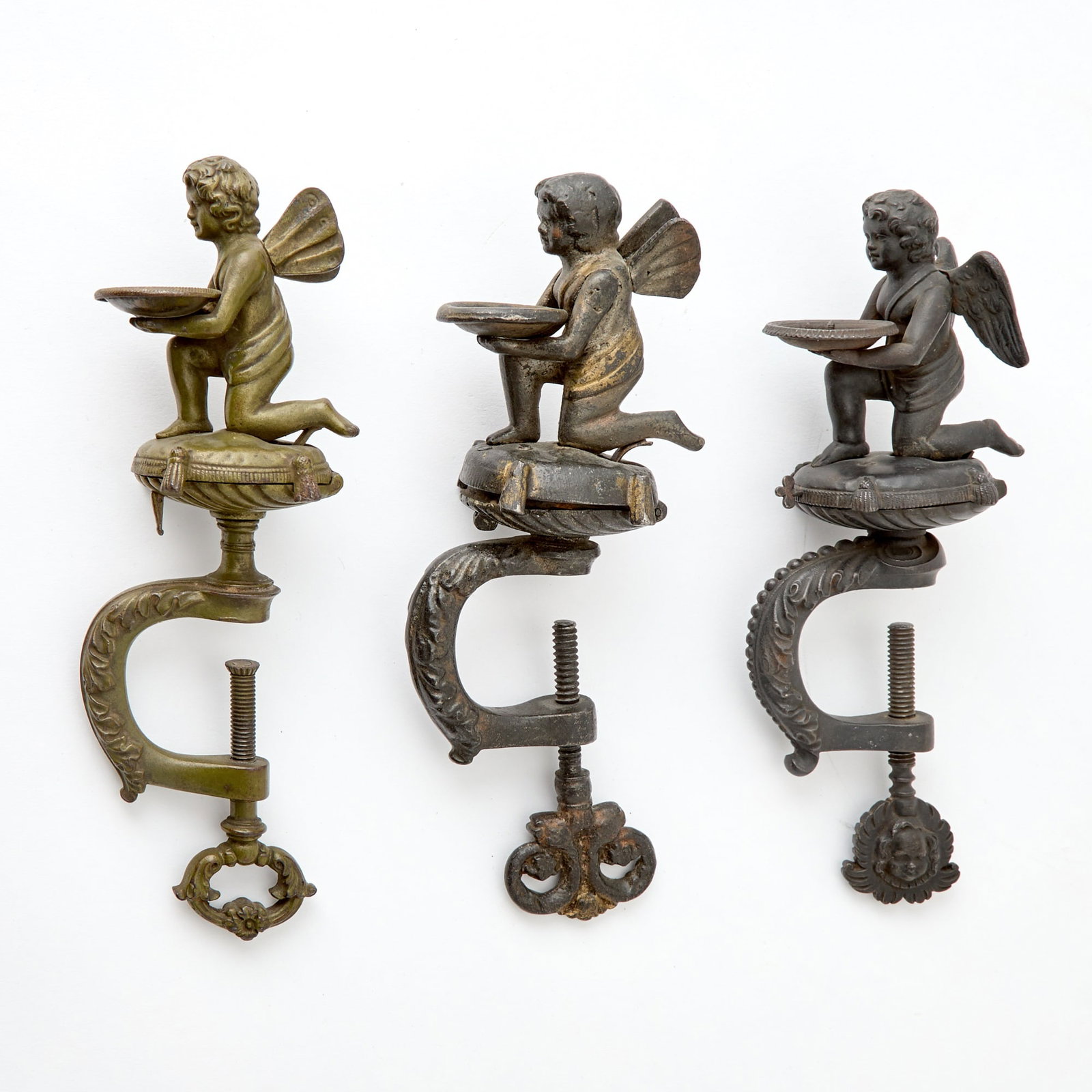 Thee Cast Iron Cupid Sewing Clamps: Second half 19th century Each cupid holding a plate for pin cushions, cushions open to reveal a storage for thread. Height of largest 8 inches.