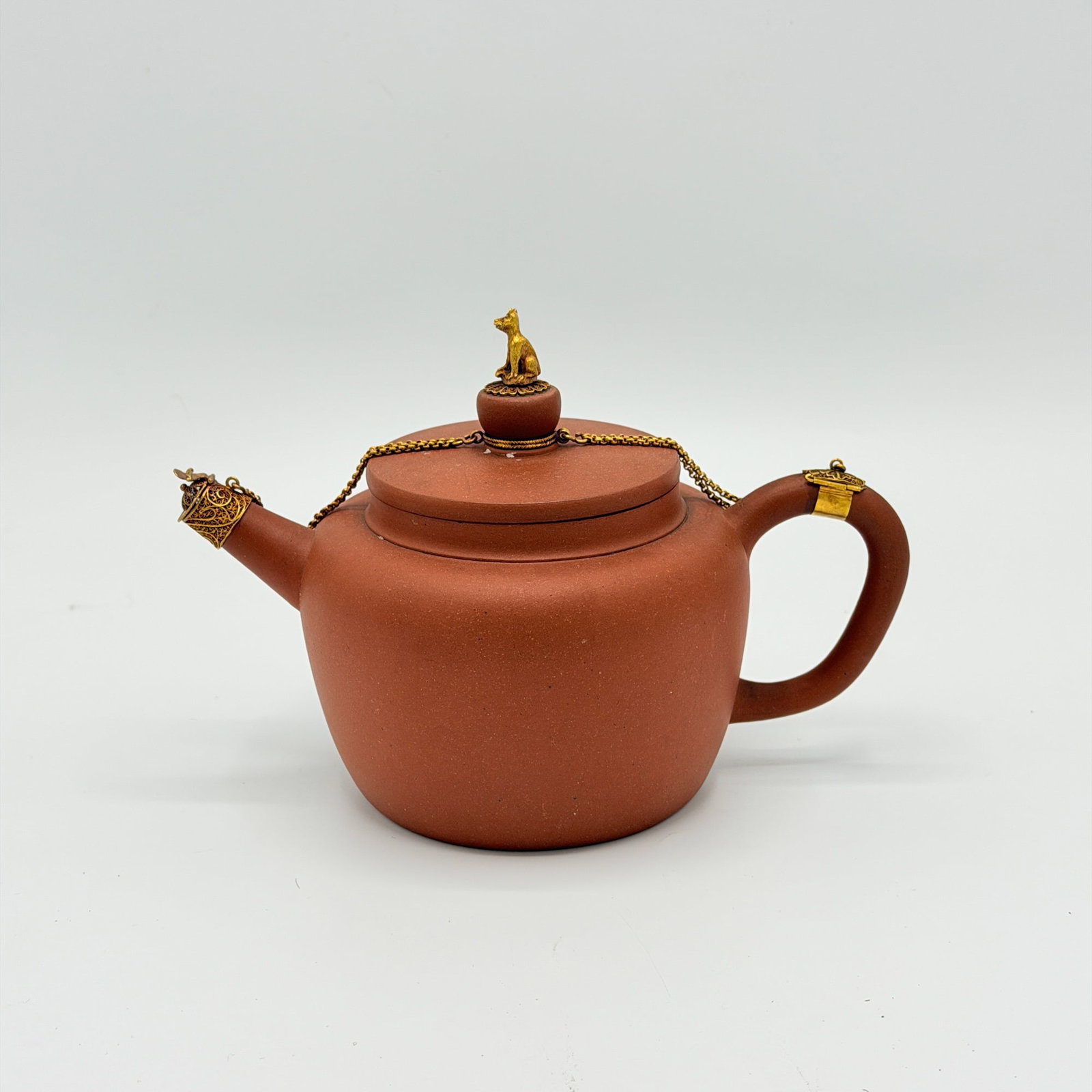 Staffordshire Redware Gilt-Metal Mounted Teapot: England, late 18th century The lid with gilt metal dog finial, attached by chains to the handle and the spout, on an unadorned body. Height 5 1/4 inches.