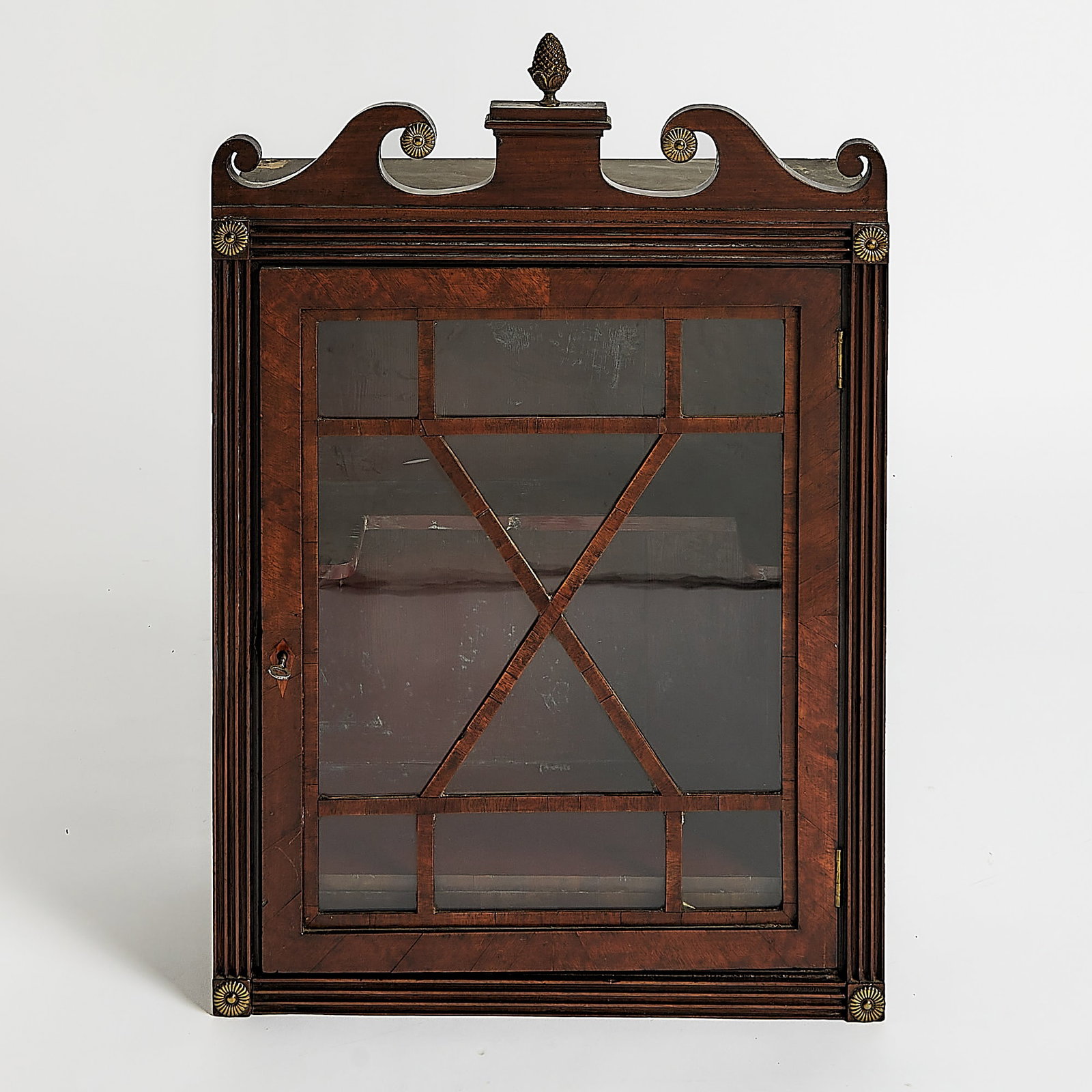 George III Mahogany Hanging Glazed Cabinet: Late 18th Century With swan's-neck cresting flanking a plinth embellished with brass rosettes and a pineapple over a glazed door enclosing shelves and a drawer within a fluted framed case. Height 29