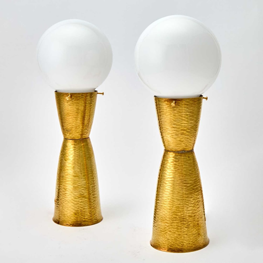 Pair of Mid-Century Modern Style Brass Table Lamps: 1960s/70s Each of hourglass form, with a textured hammered finsih and associated opaque white glass globe shade. Height overall 15 1/2 inches; height of body 10 1/4 inches.