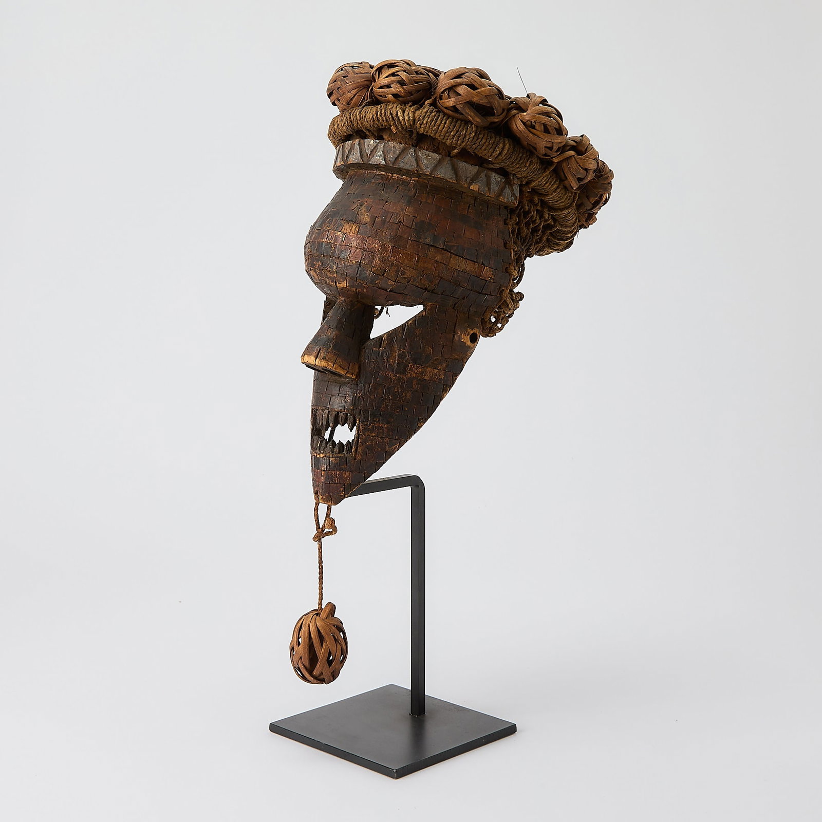 African Salampaso Carved Wood Mask: After 1960 The angular wood carved warriors mask with copper lattice over a wood core and the cane headgear woven with wicker balls at the top and as a beard. Height 13 inches.