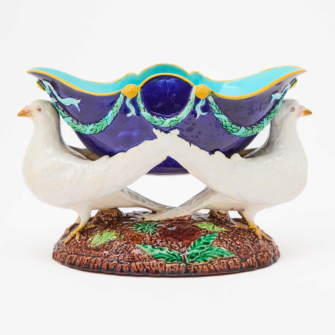 George Jones Majolica Centerpiece Bowl: Of deep oval form, the bicolored bowl with a turquoise interior, cobalt exterior, and yellow rim, decorated with garland swags, raised on two opposed white doves, ending in a naturalistic domed foot. 
