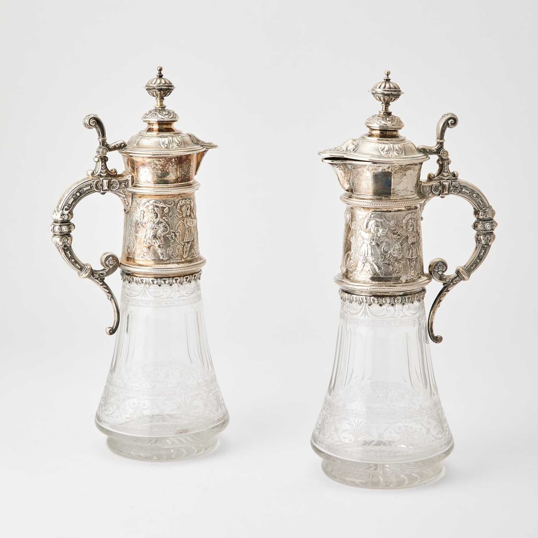 Pair of German Silver Mounted Etched Glass Claret Jugs: Retailed by Gebruder Friedlander, late 19th/early 20th century The faceted spreading cylindrical body with silver neck chased with figures and foliate scrolls, the hinged covers with button finials. H