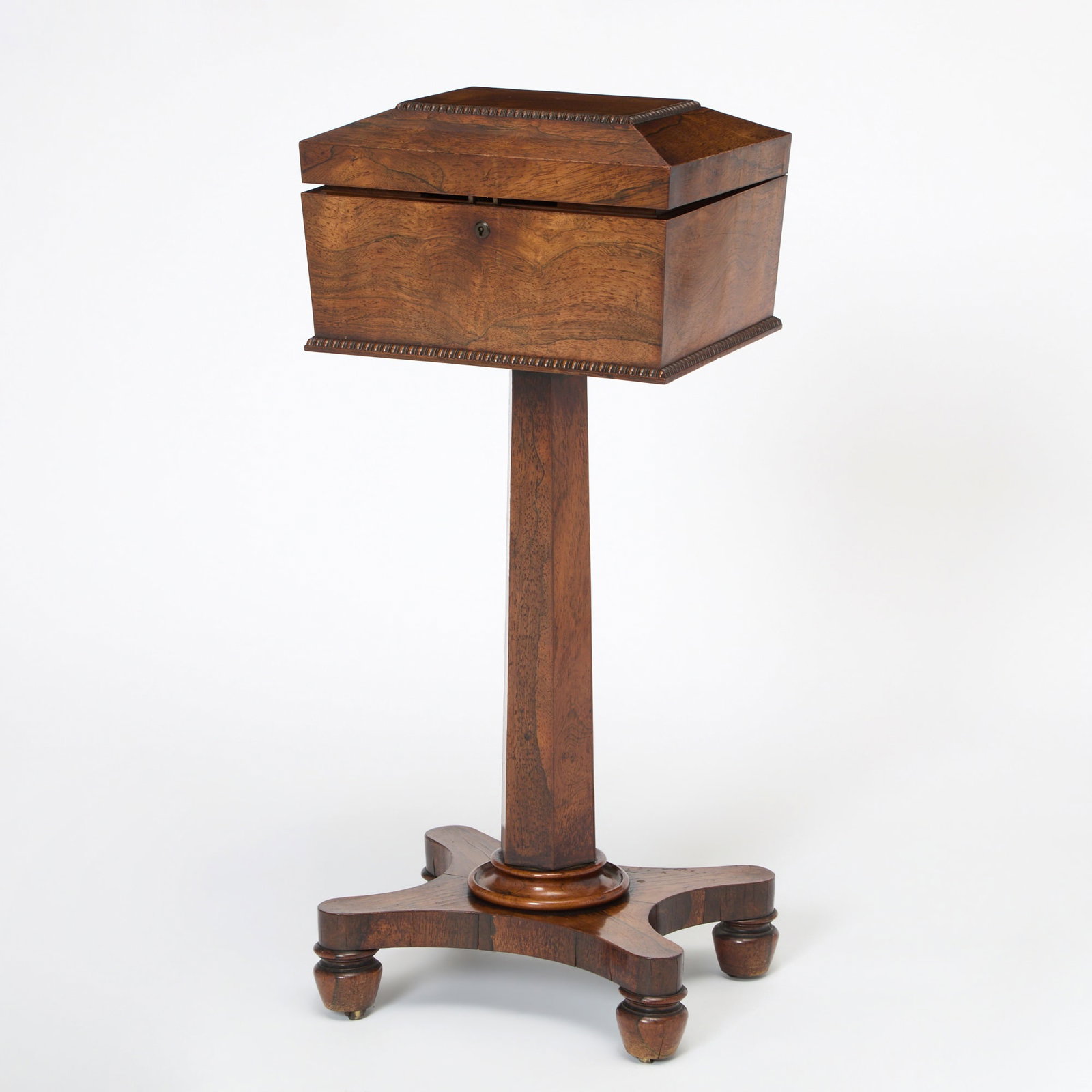 Regency Rosewood Teapoy: Circa 1815 The hinged rectangular top with beaded moldings, opening to an interior with covered compartments and two cut-glass bowls, raised on a hexagonal stem over a quadripartite base and ending