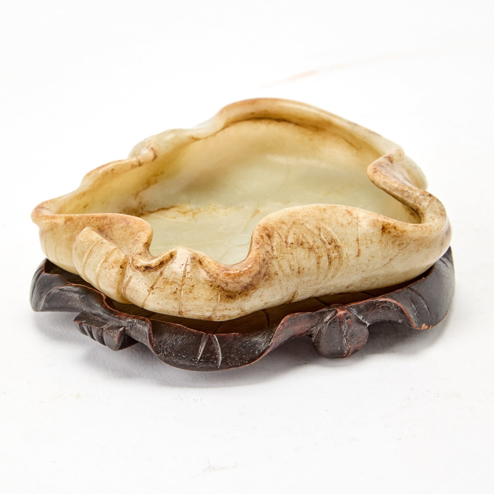 A Chinese Russet and Pale Celadon Jade Carving Qing Dynasty: Carved as a lotus leaf with the edges curled upward, the underside carved with stems and lotus blossoms; with a fitted wood stand. (Length 4 1/4 inches)
