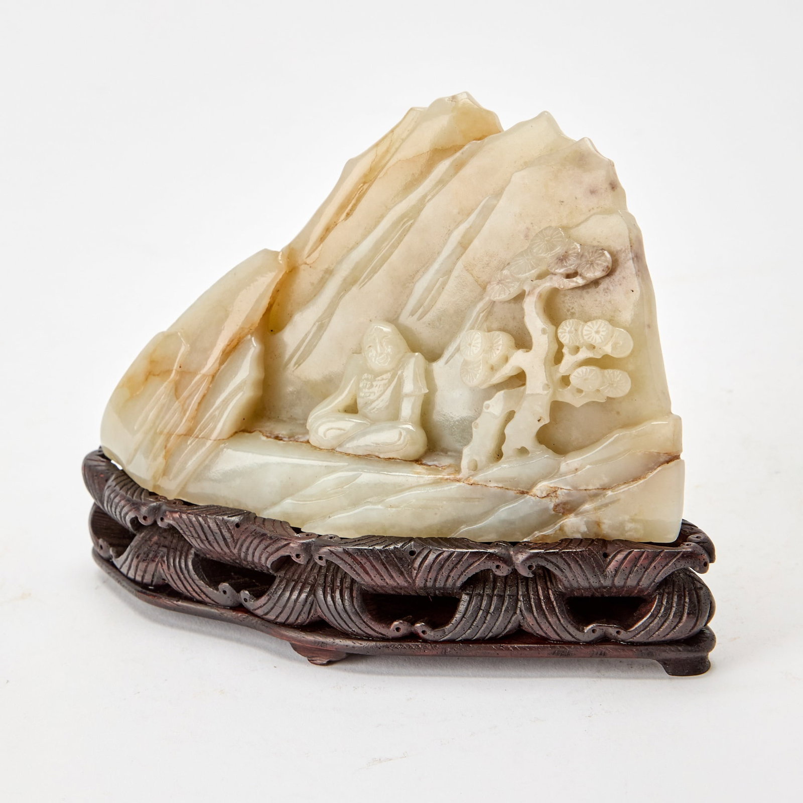 A Chinese Russet and Celadon Jade Boulder Qing Dynasty: Carved with a figure seated beneath a pine tree, with mountains in the background; with a fitted wood stand. (Length 5 3/4 inches)
