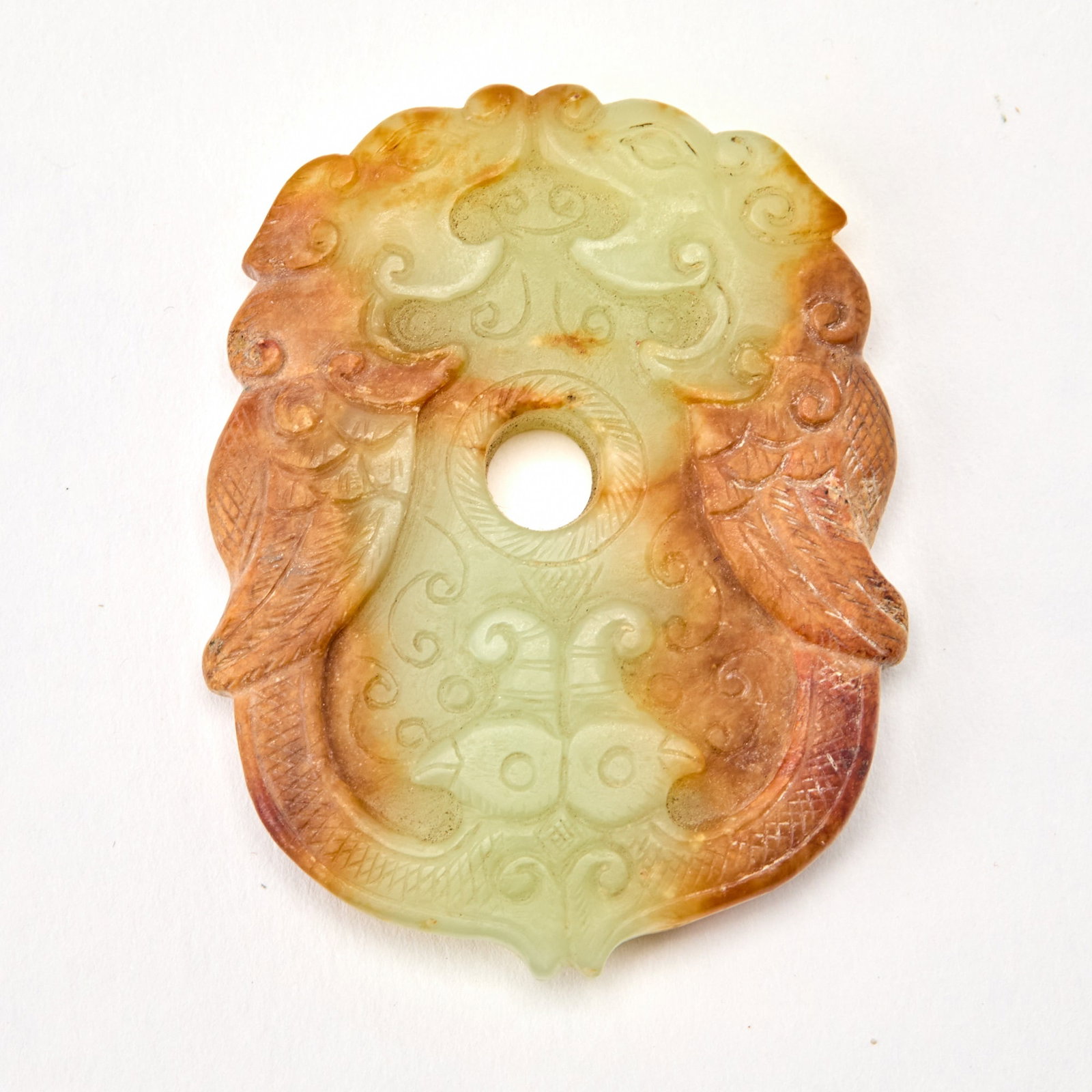 A Chinese Carved Celadon and Russet Jade Pendant Qing Dynasty: Decorated with a pair of confronting phoenix encircling a central aperture. (Length 2 inches)