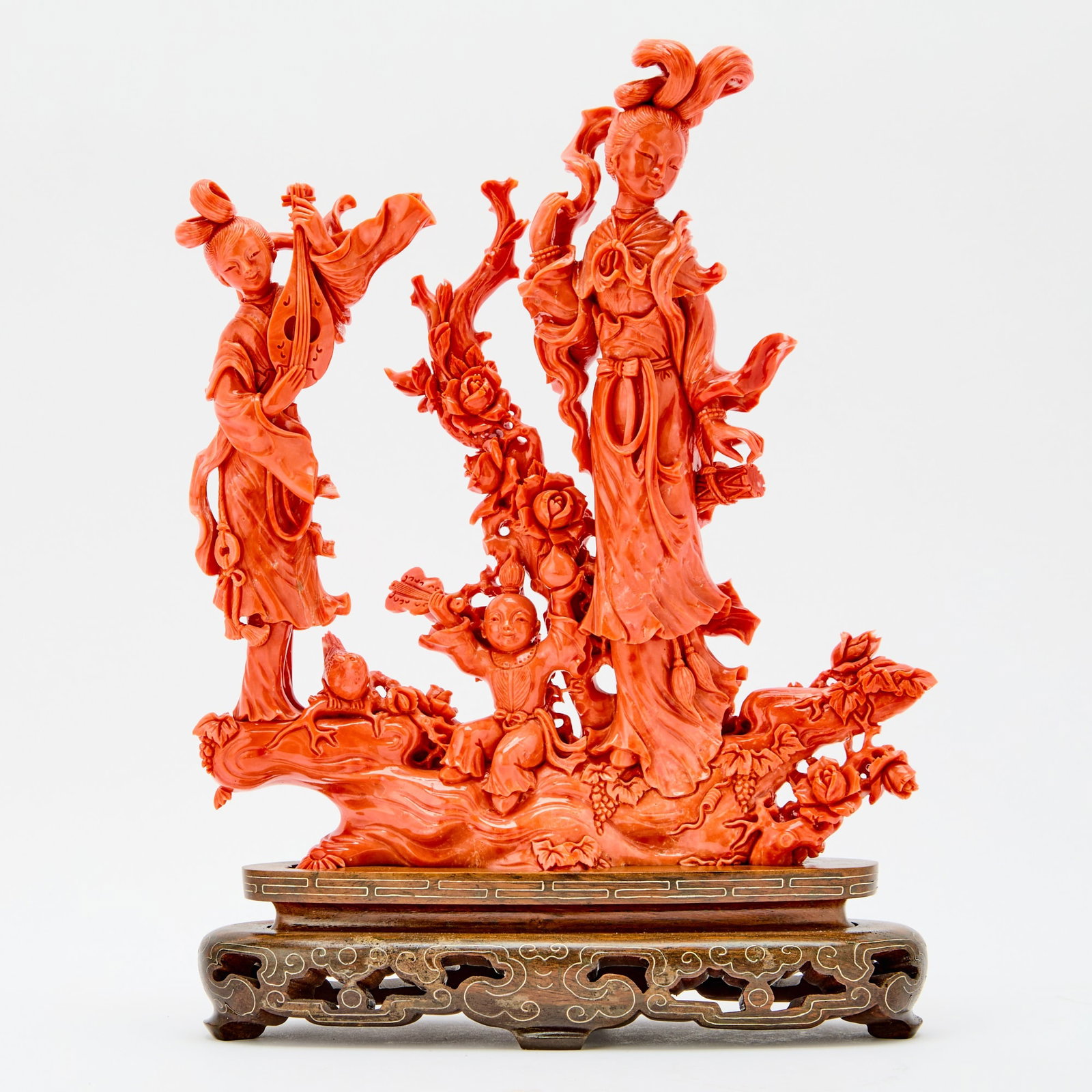 A Chinese Coral Figural Group Carving: 20th Century Carved with two meiren dressed in long flowing robes and wearing high chignons, one playing the pipa, accompanied by a boy holding a peach, all standing on rockwork decorated with leafy a