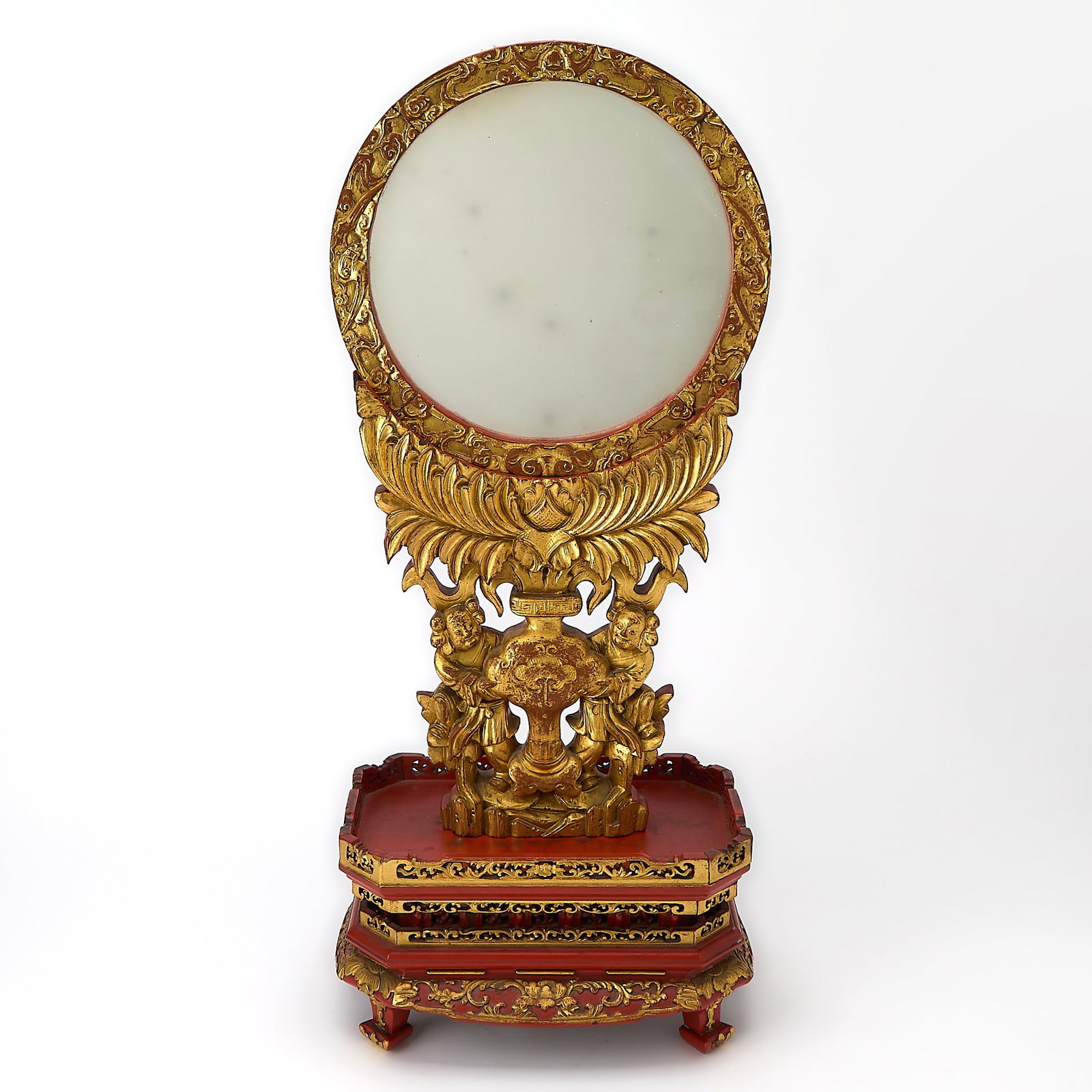 A Chinese Giltwood Mirrored Table Screen 19th Century: The waisted pedestal supported on four legs, the sides reticulately carved with lotus scrolls, surmounted by two attendants flanking a vase issuing a large blossom that supports the circular mirror.
