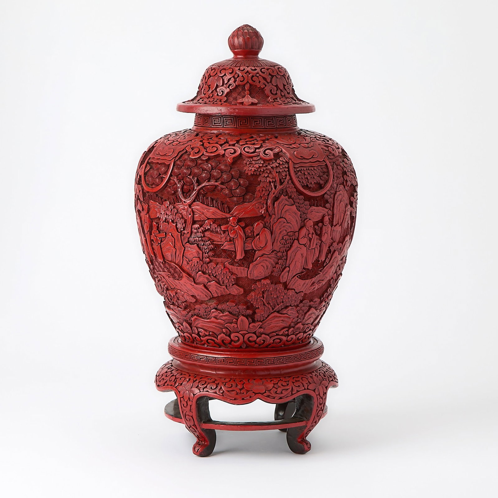 A Chinese Carved Cinnabar Lacquer Vase and Stand Republic Period: The vase carved to depict scholars and attendants amidst a lush mountainous landscape, set on a stand raised on four cabriole legs and surmounted by a domed cover with a pointed finial. (Height 12 inc