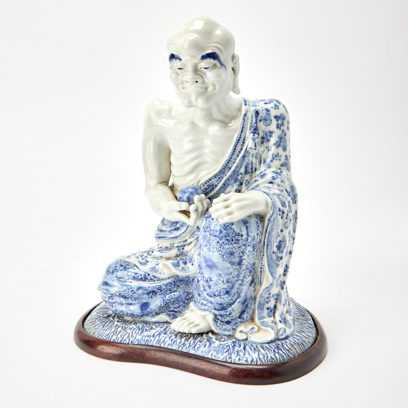 A Chinese Blue and White Porcelain Figure of a Luohan Republic Period: Modeled as a seated luohan with a gaunt torso, bald head, and arched brows, wearing loosely draped robes painted with floral and foliate designs, his right hand holding a ball. (Height 12 1/2 inches) 