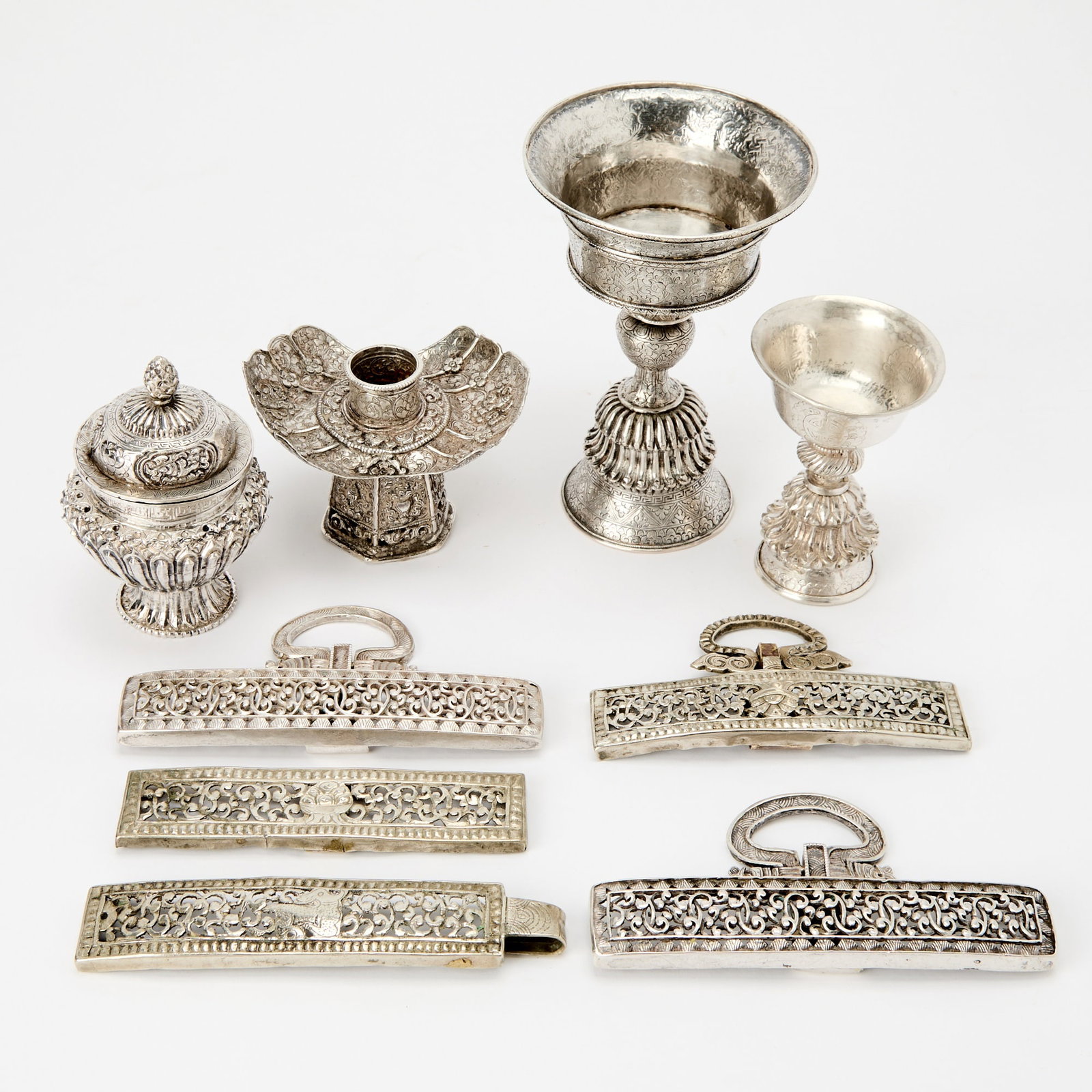 A Group of Tibetan Silver and Silvered Metal Ritual Articles Likely 19th century: Comprising a large and a small joined two-piece buttter lamp, footed jar and domed cover, a repousse candle holder with auspicious emblems, and five reticulated belt plaques including a pair of fine w